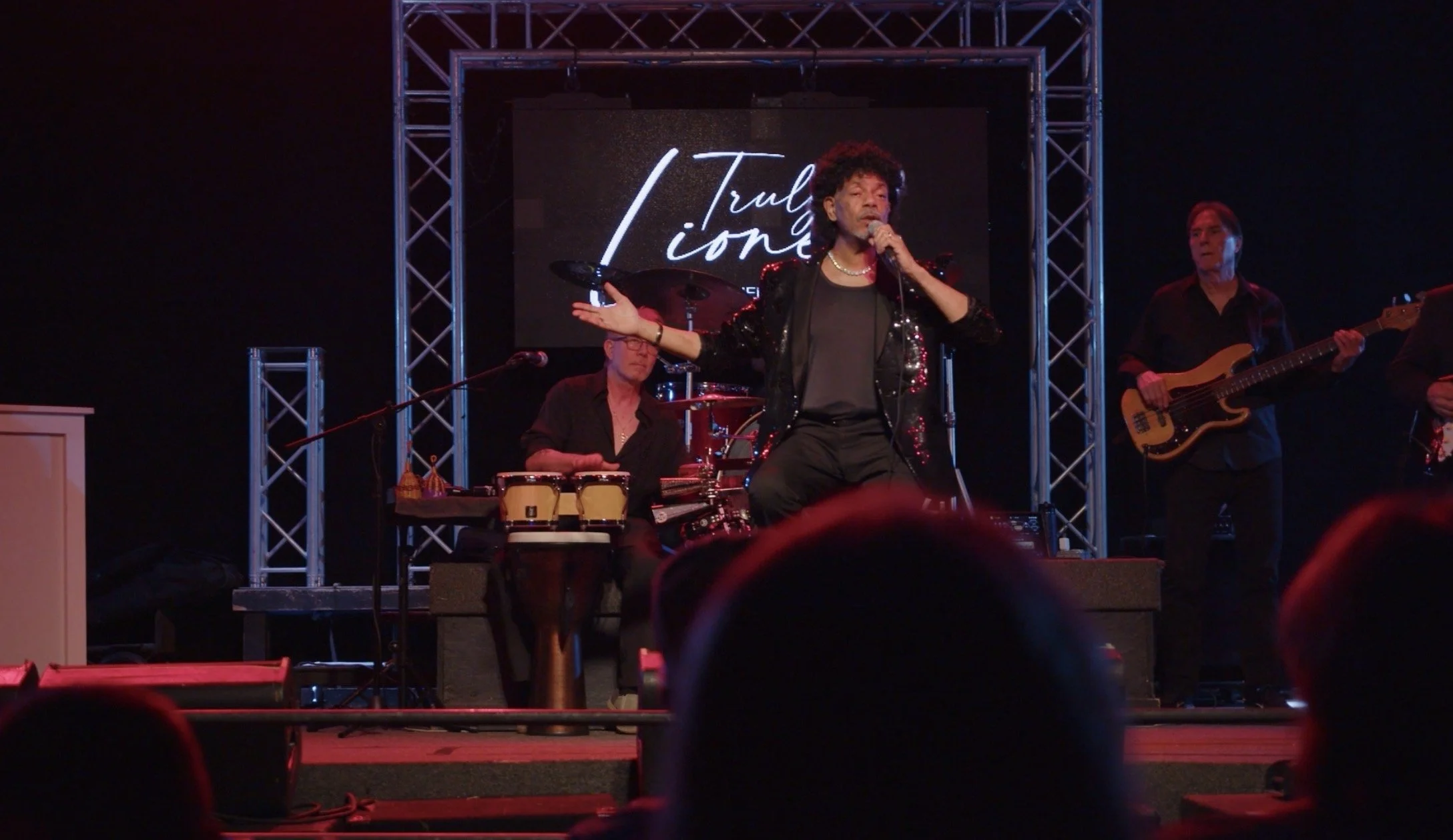 A person singing on stage with a microphone, dressed in black with a sequined jacket, flanked by musicians playing instruments, with a backdrop displaying the words Truly Lionel: Lionel Richie Tribute