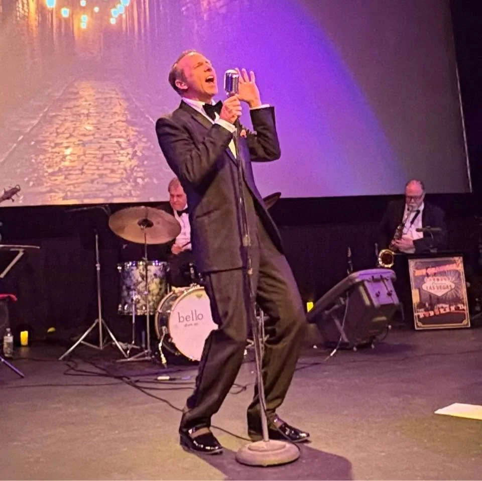 A man in a tuxedo singing into a vintage microphone on stage with a band, including a drummer and saxophonist, during a performance.