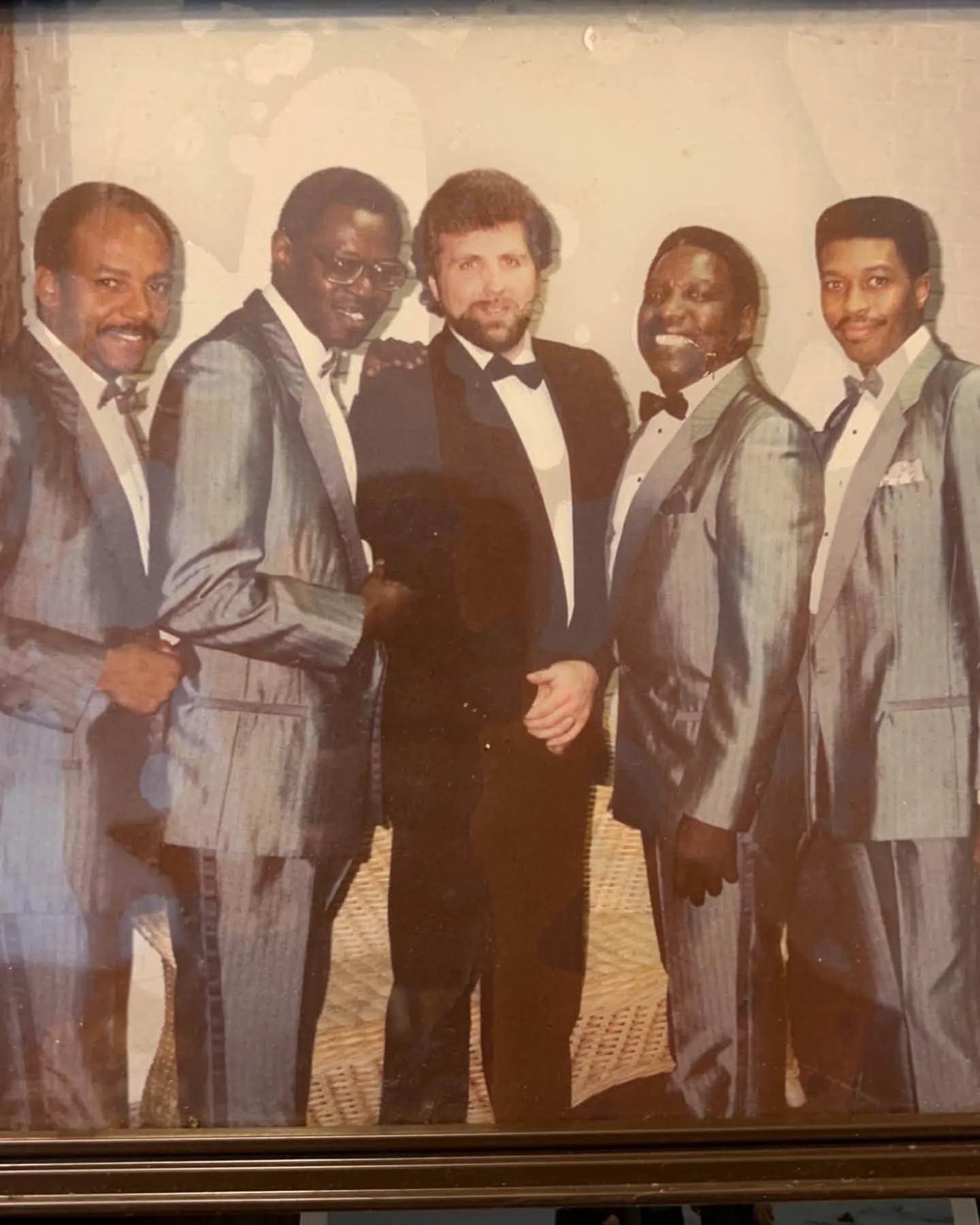 A group of six men dressed in tuxedos, standing together indoors. They are smiling and posing for the photo. The background is a plain wall, and the image has a vintage or old photograph appearance.