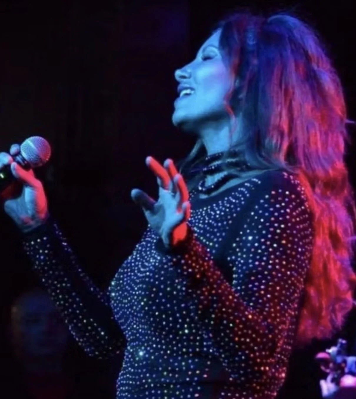 Performer singing or speaking into a microphone on stage, wearing a black outfit adorned with small shiny embellishments, with colorful stage lighting illuminating her face and hair.