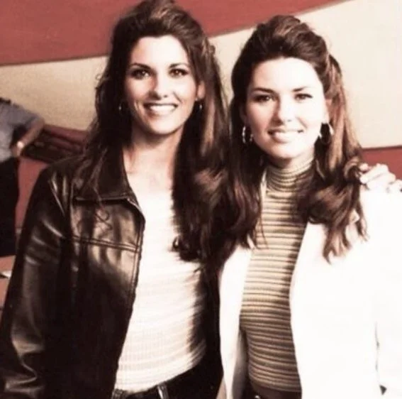 Two women with long, wavy brown hair smiling at the camera, one wearing a black leather jacket and the other in a white blazer over a striped turtleneck, standing close together indoors.