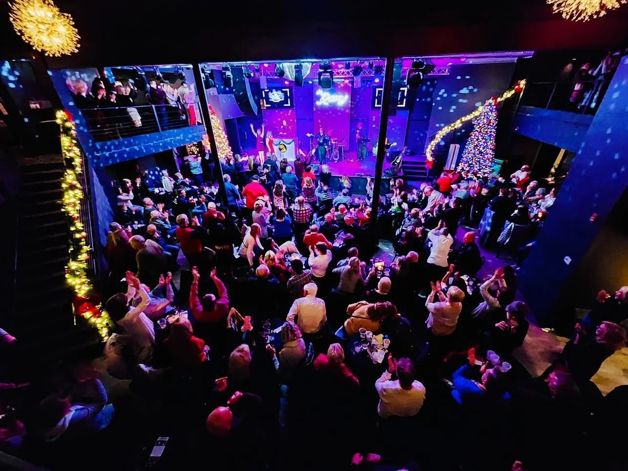 A lively indoor holiday concert with an audience, stage lighting, Christmas trees, and a band performing on a stage decorated with festive lights.