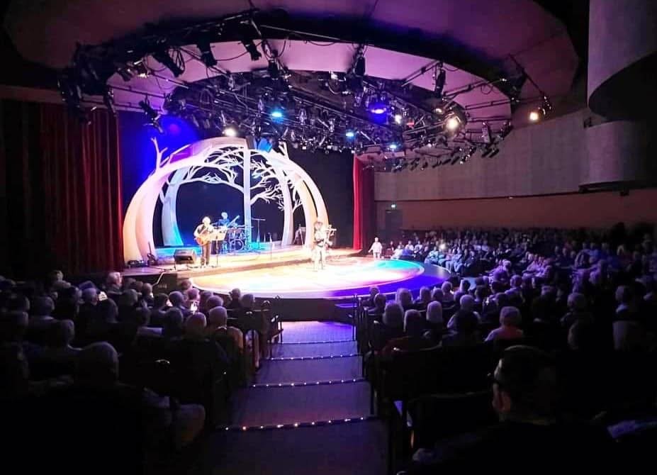 A theater stage with a musical performance in front of an audience. The stage has a circular, forest-themed set with arching tree designs, and musicians are playing guitars with a drummer in the background. Audience members sit in dark theater seats watching the show.