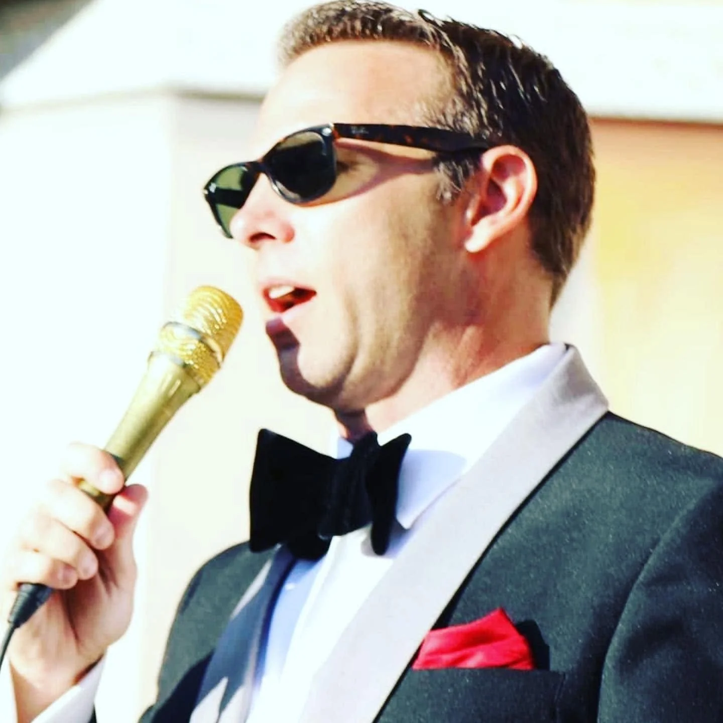 Man wearing tuxedo, bow tie, and sunglasses speaking into a gold microphone.