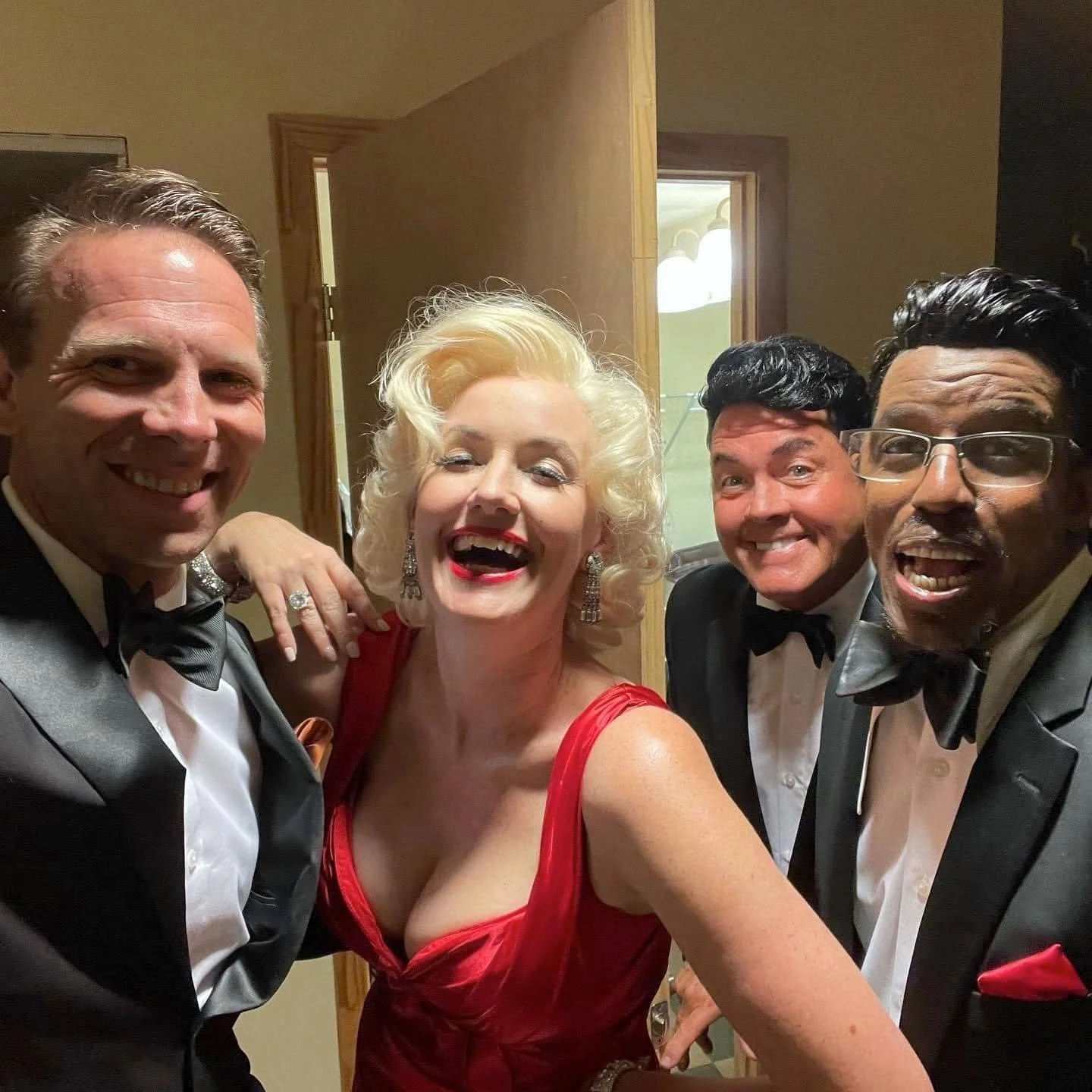 Four people in formal tuxedos and a woman in a red dress taking a selfie together at an event, all smiling.