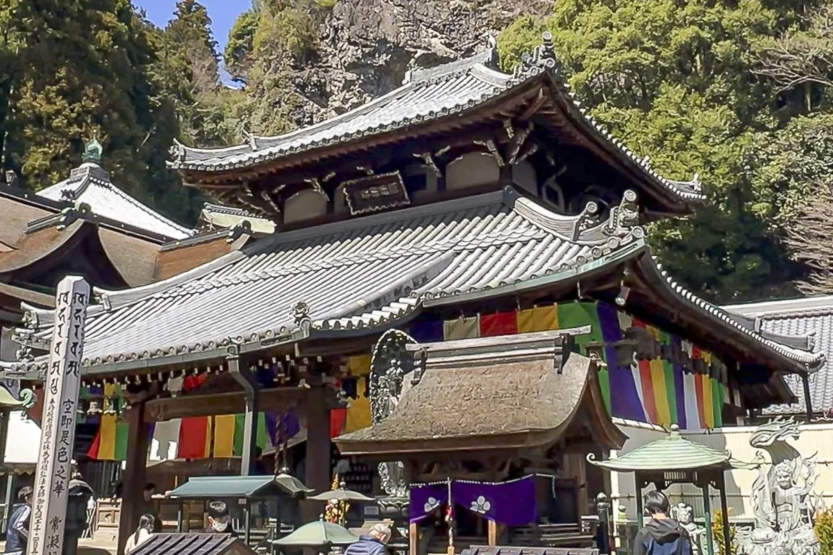 Traditional Japanese temple with multi-tiered curved roof, colorful Buddhist banners, and stone statues, set against lush green trees.