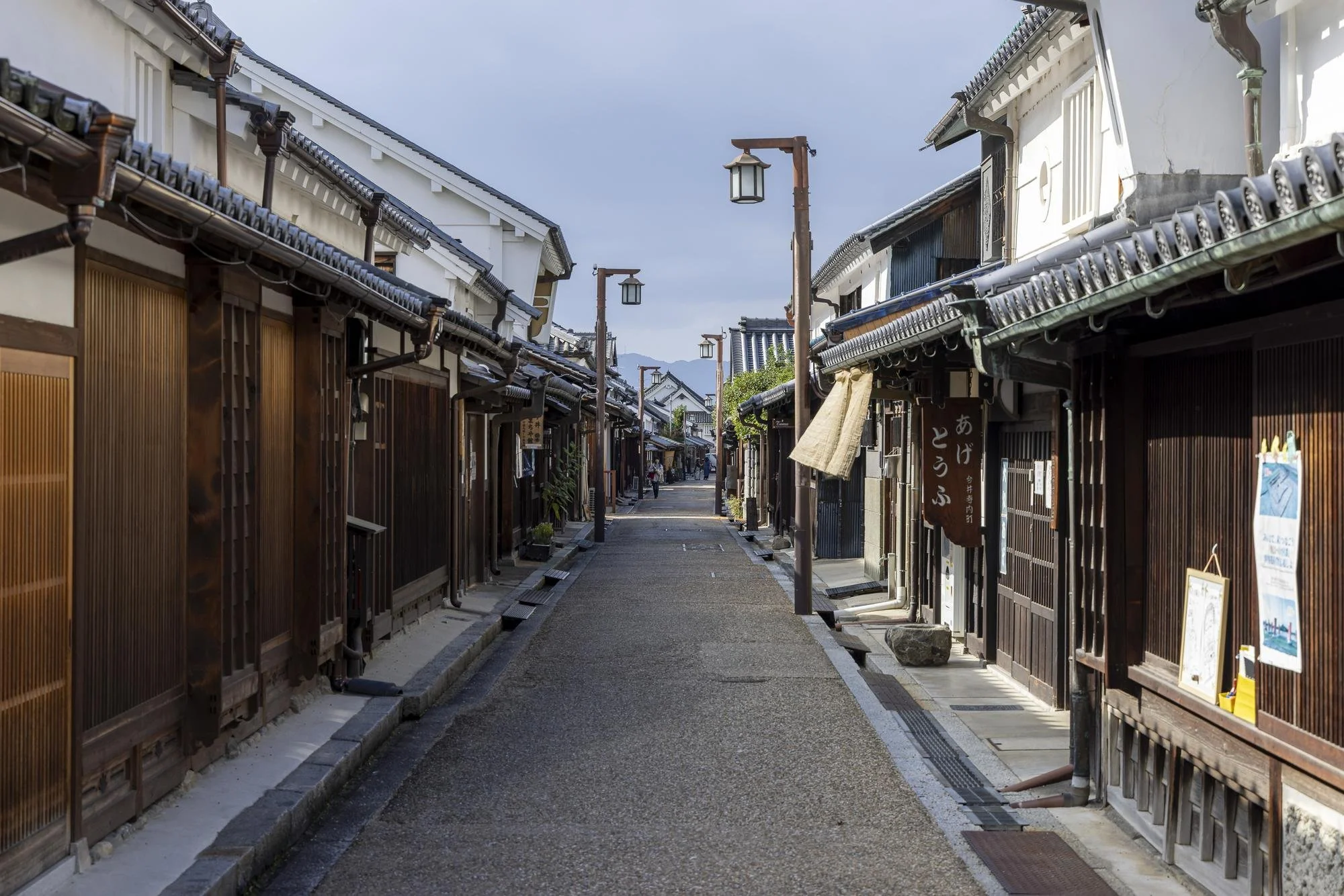 A quiet historical street in Japan with traditional wooden buildings, narrow pathway, and stone lanterns, under an overcast sky.