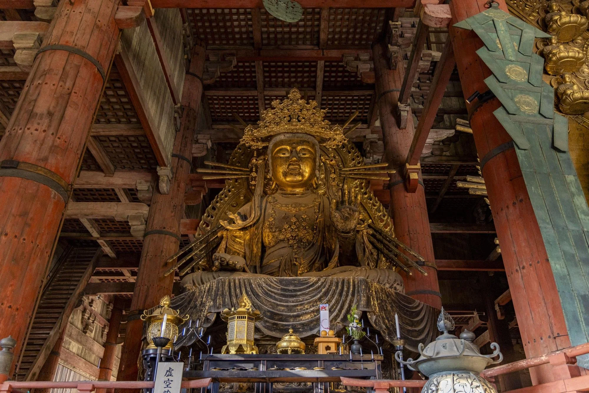 Large, gold-colored statue of a seated figure inside a traditional Japanese temple, surrounded by wooden pillars and ornate decorations, with offerings placed in front.