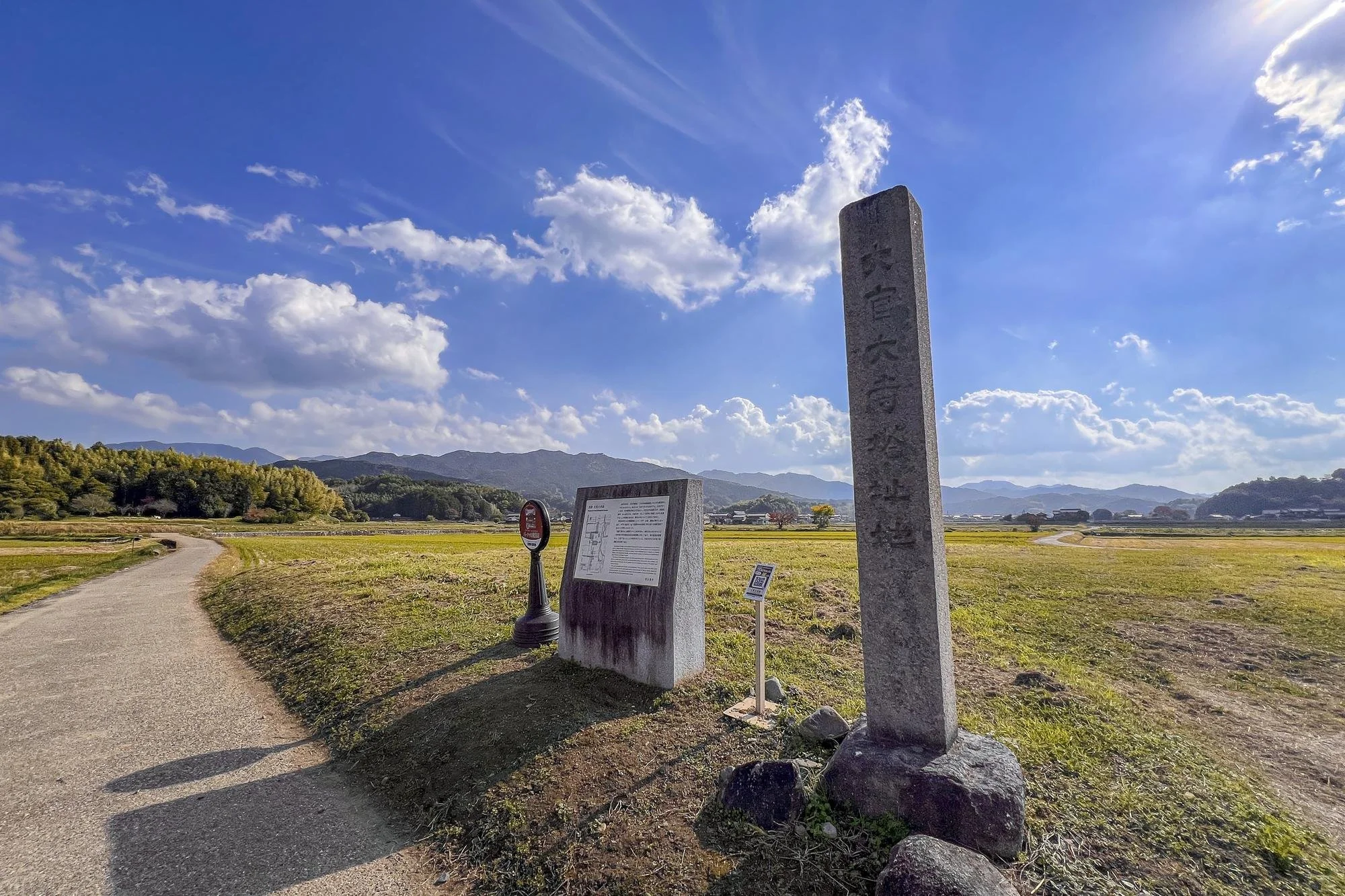 Open field with a winding dirt path, standing signs, and a scenic mountain range in the background under a partly cloudy blue sky.