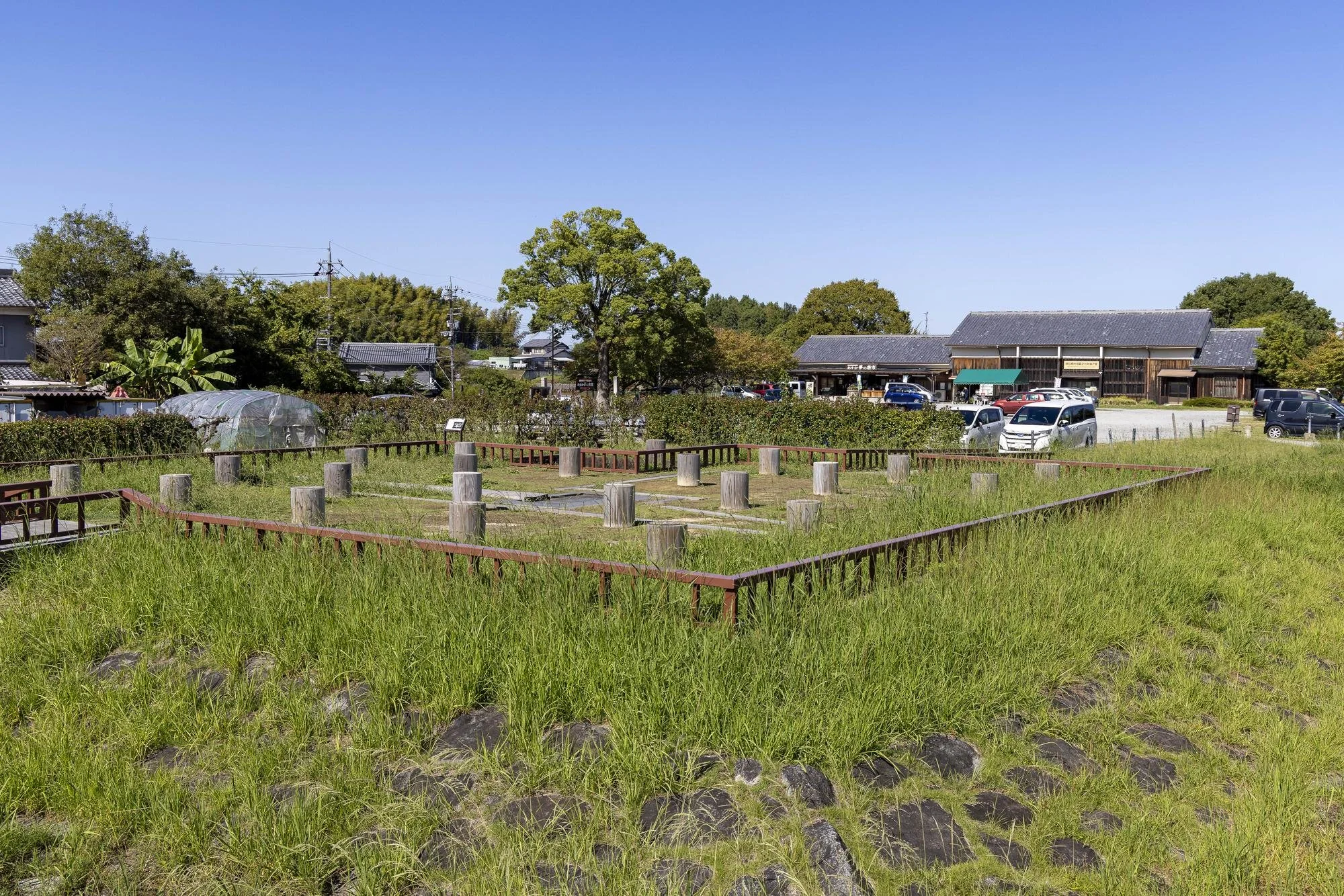 Open grassy field with wooden posts and railing outline, surrounded by tall green grass and trees, with buildings and parked cars in the background under a clear blue sky.