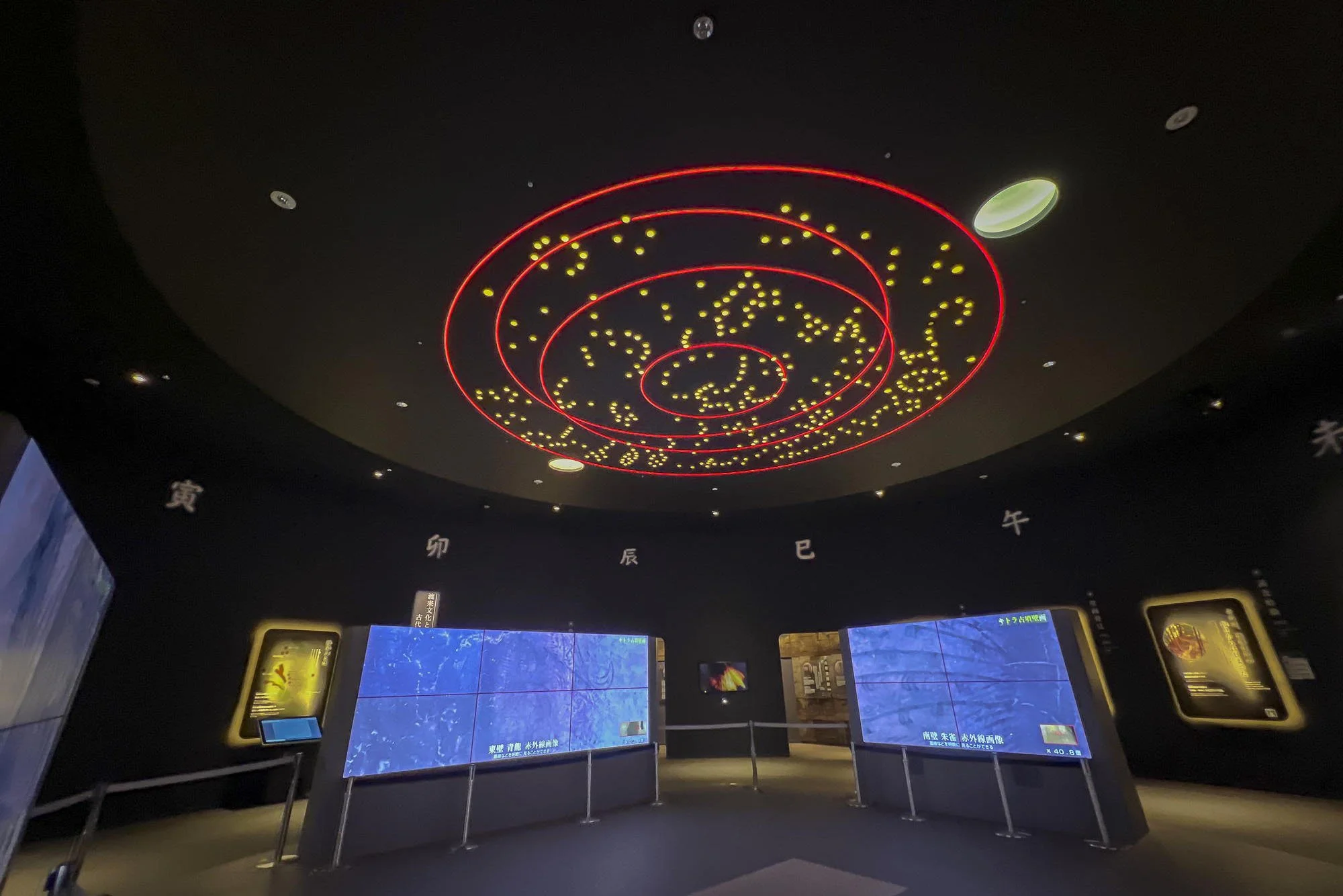 Exhibit inside a museum showing a black ceiling with illuminated zodiac symbols and a circular arrangement of red and yellow lights depicting a galaxy or constellation. There are digital screens on the walls displaying information.