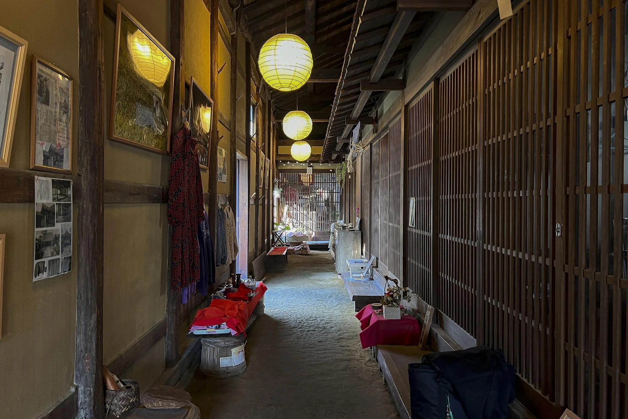 Interior view of a traditional Japanese-style room with wooden walls, hanging paper lanterns, framed pictures, and small tables with various items, leading to an open area with natural light.