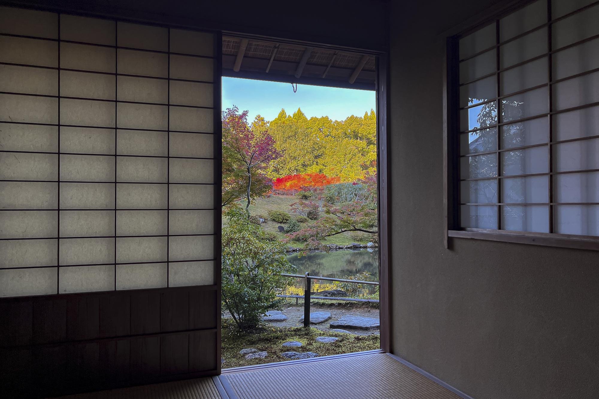Interior of a traditional Japanese room looking out to colorful autumn trees and a pond, with sliding shoji doors and tatami flooring.