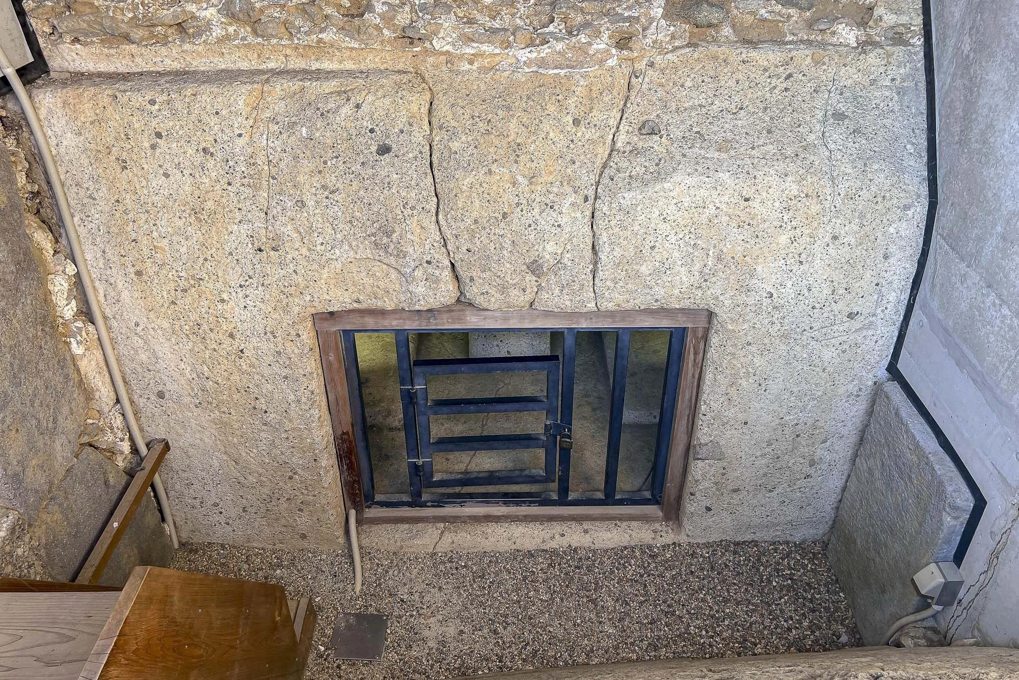 A concrete wall with cracks, a small barred window with a wooden frame, and some electrical cords and a vent on the floor.