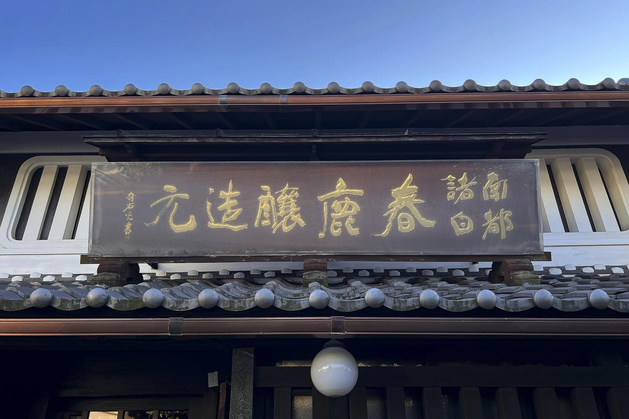 Traditional Japanese building with a wooden signboard displaying Japanese characters in gold, white decorative elements, and a tiled roof, under a clear blue sky.