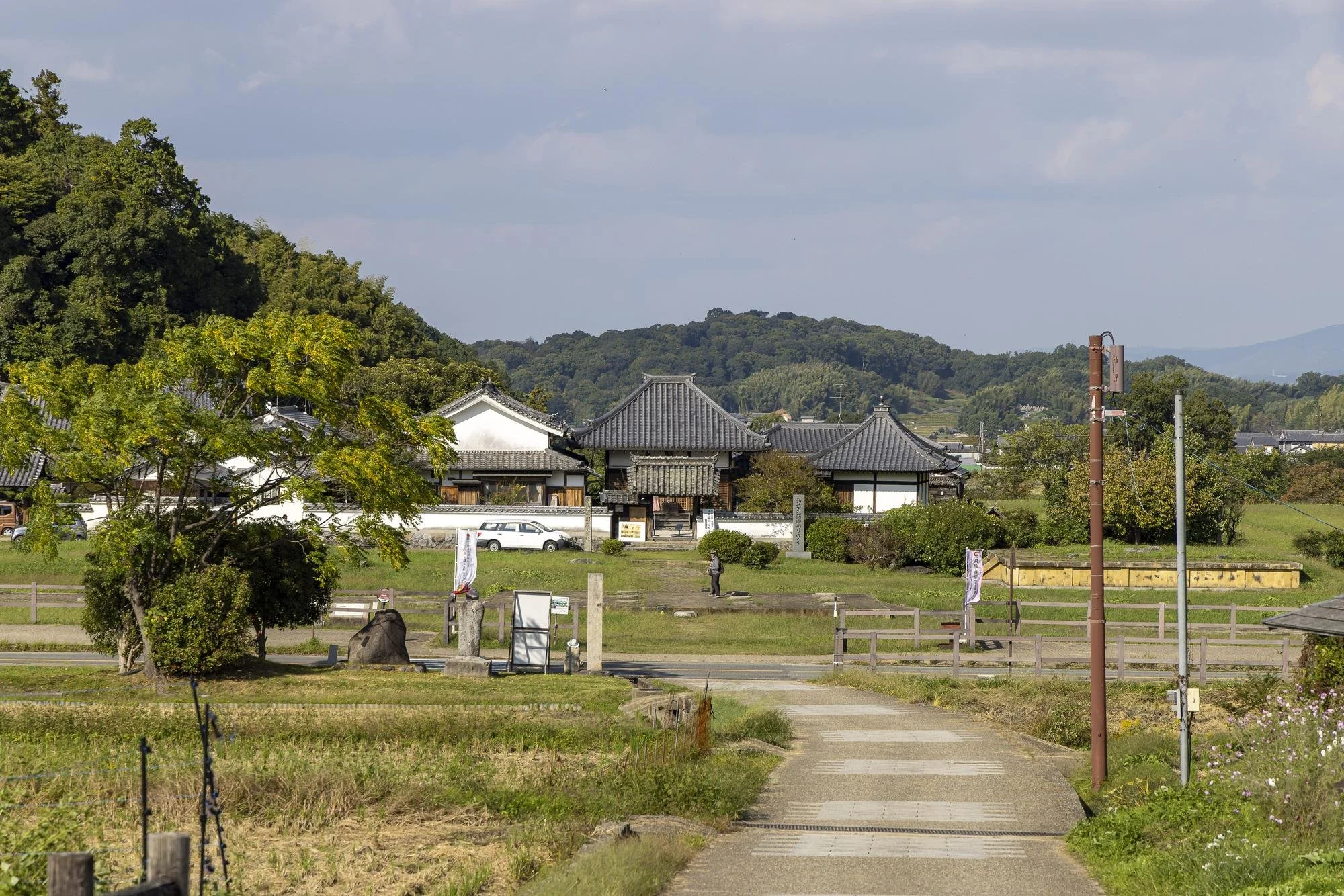 A rural scene with traditional Japanese-style buildings, a tree on the left, green hills in the background, and a pathway leading towards the buildings.