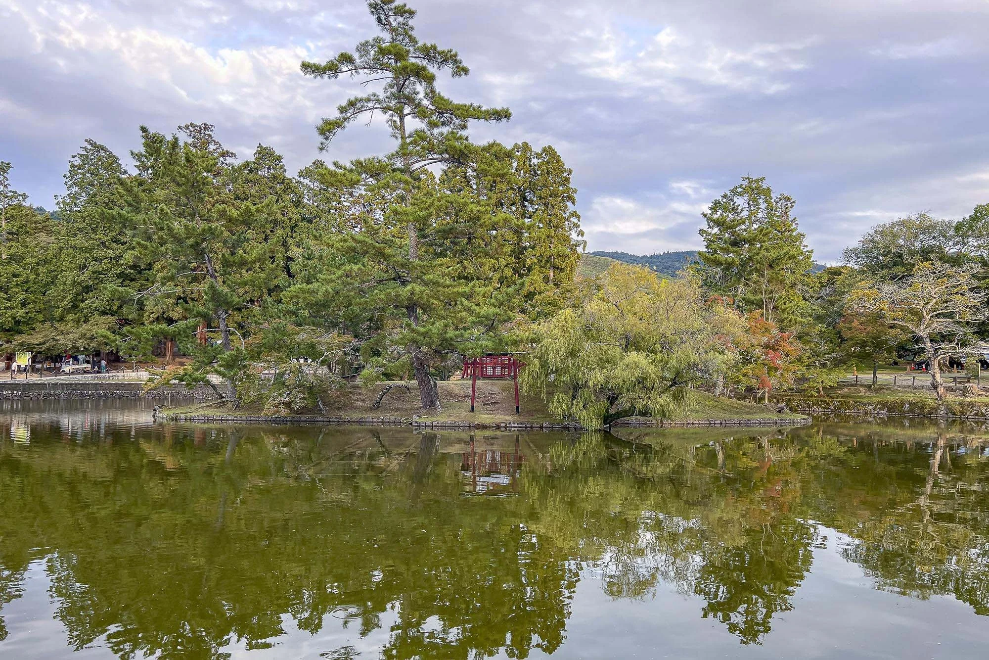 A serene landscape with a calm body of water reflecting the trees and a red torii gate on a small island. The background features a cloudy sky and distant rolling hills.