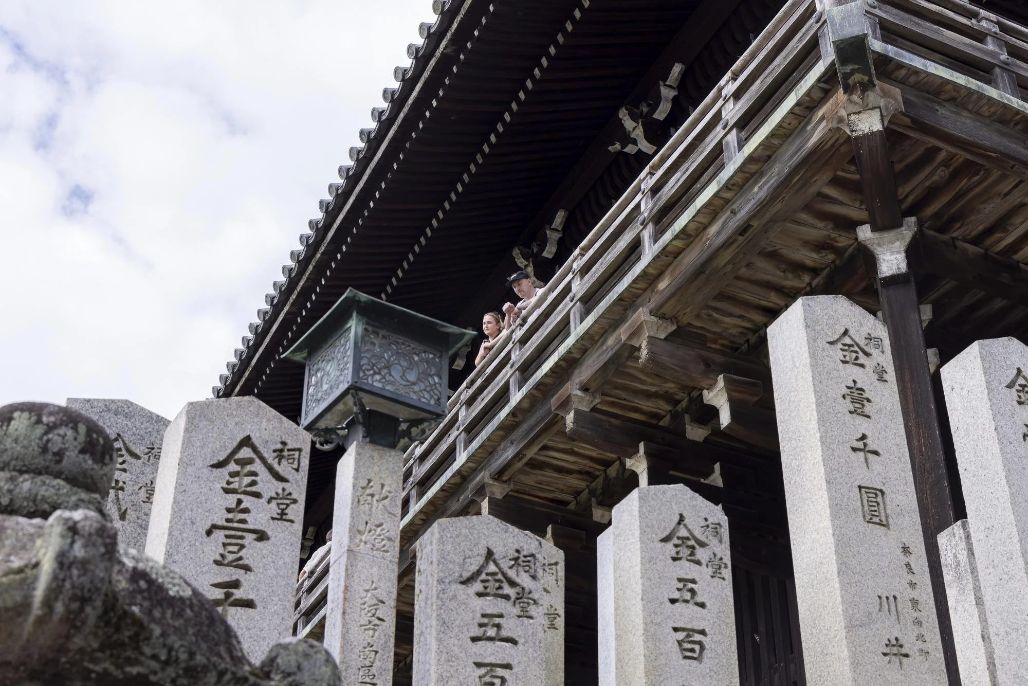 People standing on a wooden balcony of a traditional Japanese temple, with stone and engraved markers in the foreground and cloudy sky in the background.