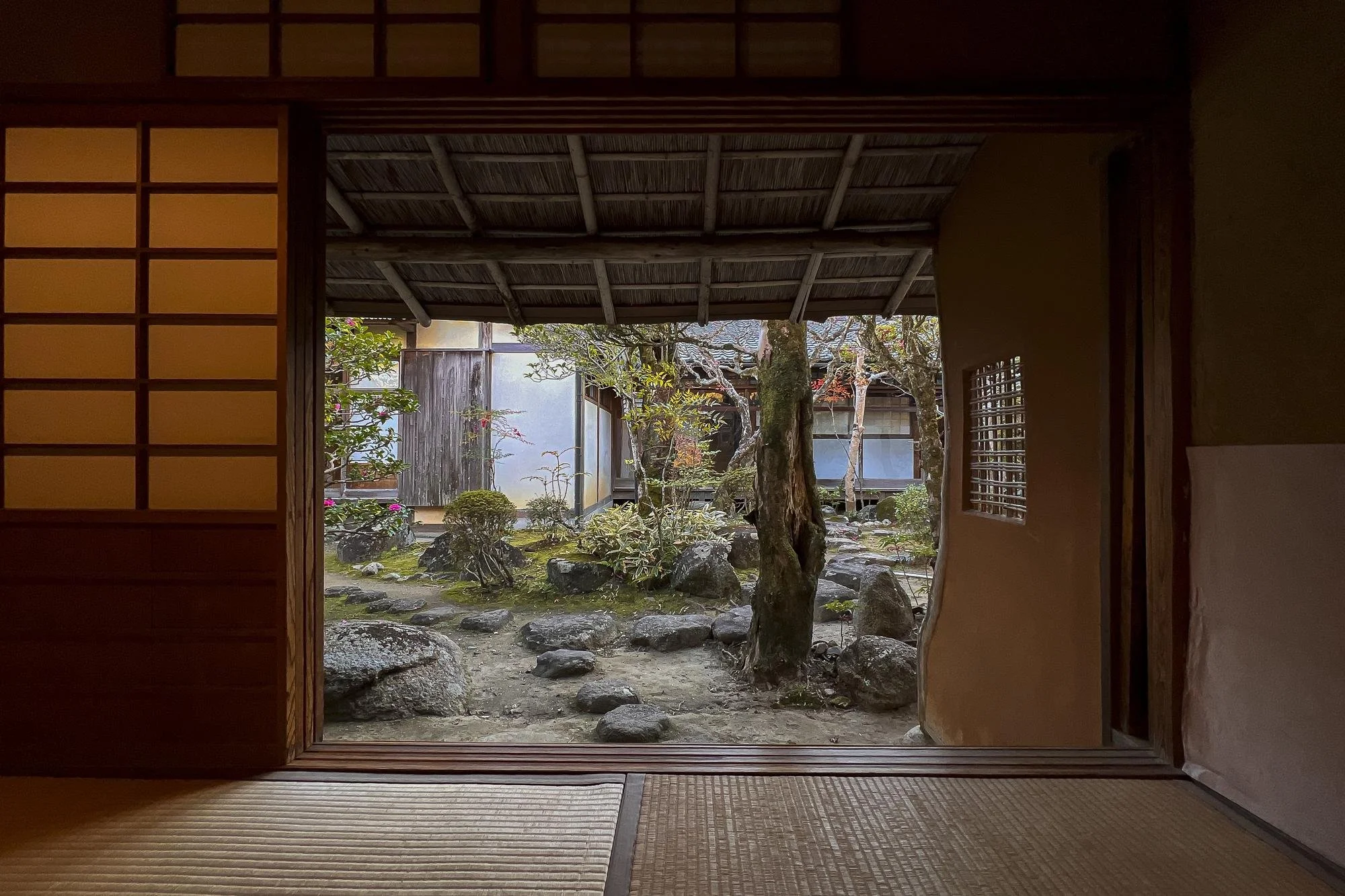Traditional Japanese room viewing a garden with rocks, bushes, and trees through an open sliding door.