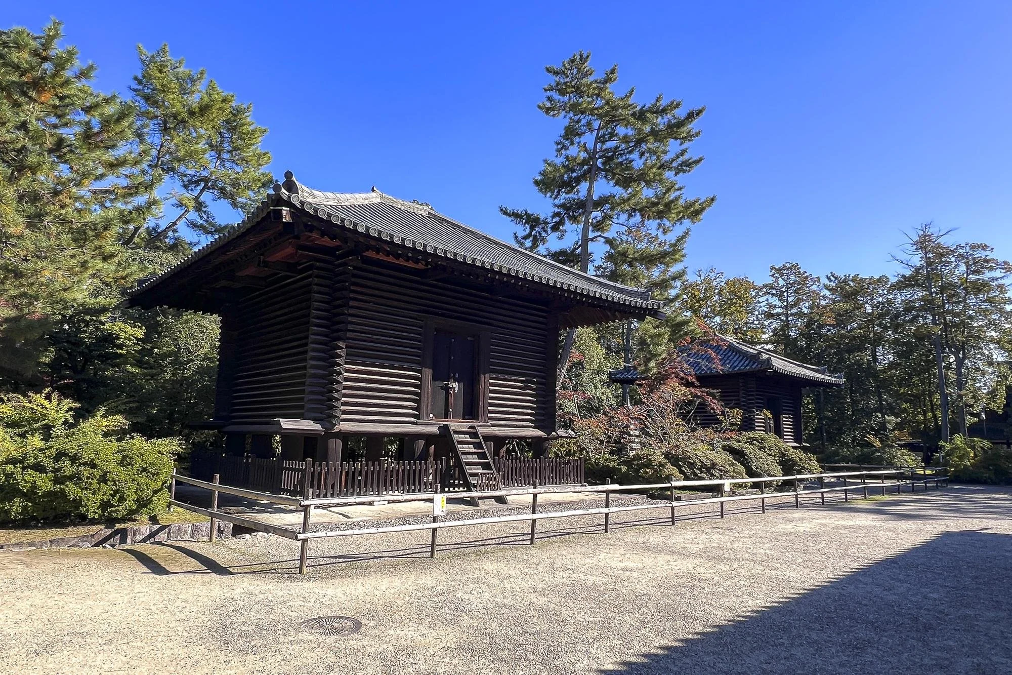 Traditional Japanese wooden buildings in a garden surrounded by trees and greenery under a clear blue sky.