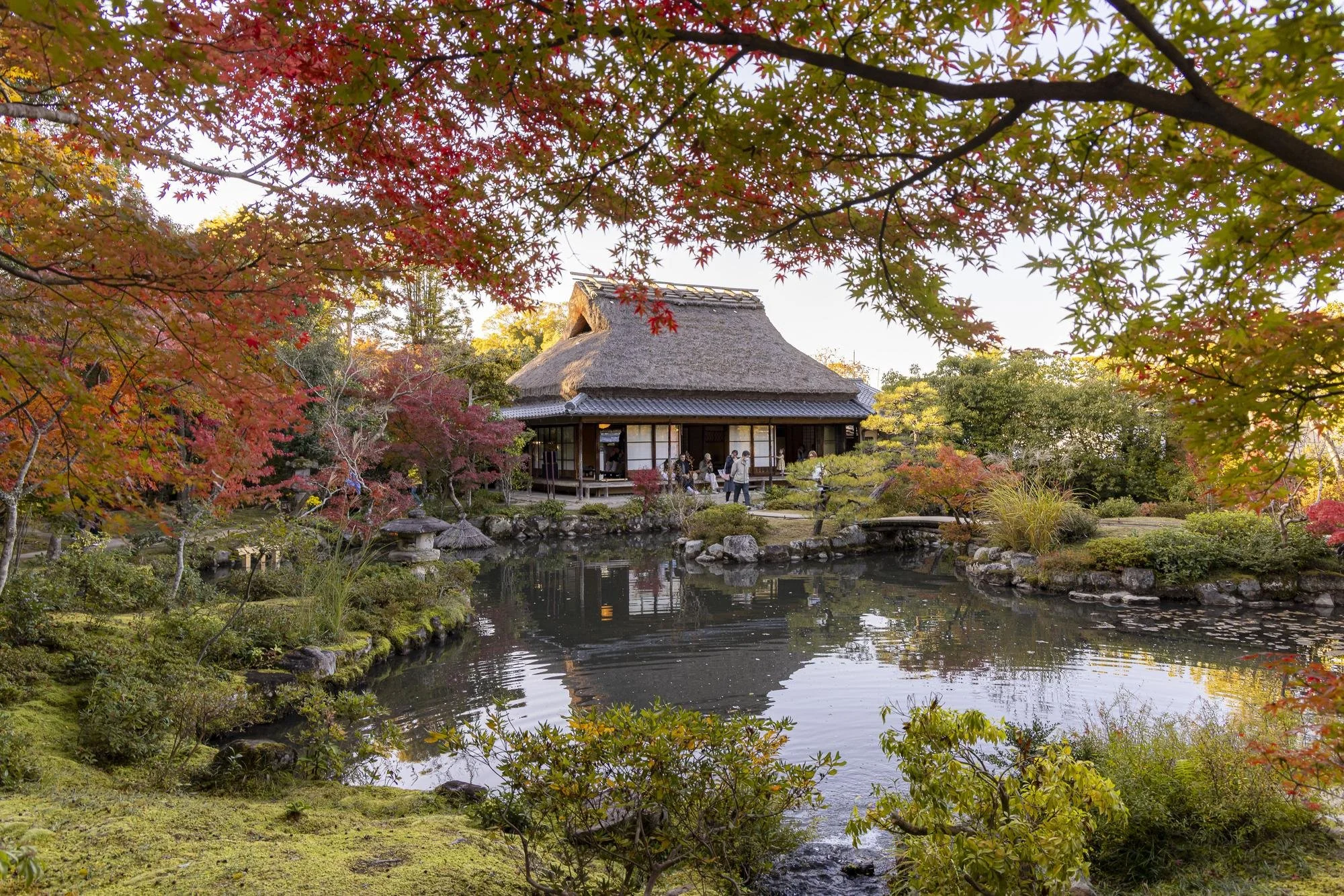 A traditional Japanese garden with a pond, colorful autumn trees, and a thatched-roof house in the background.