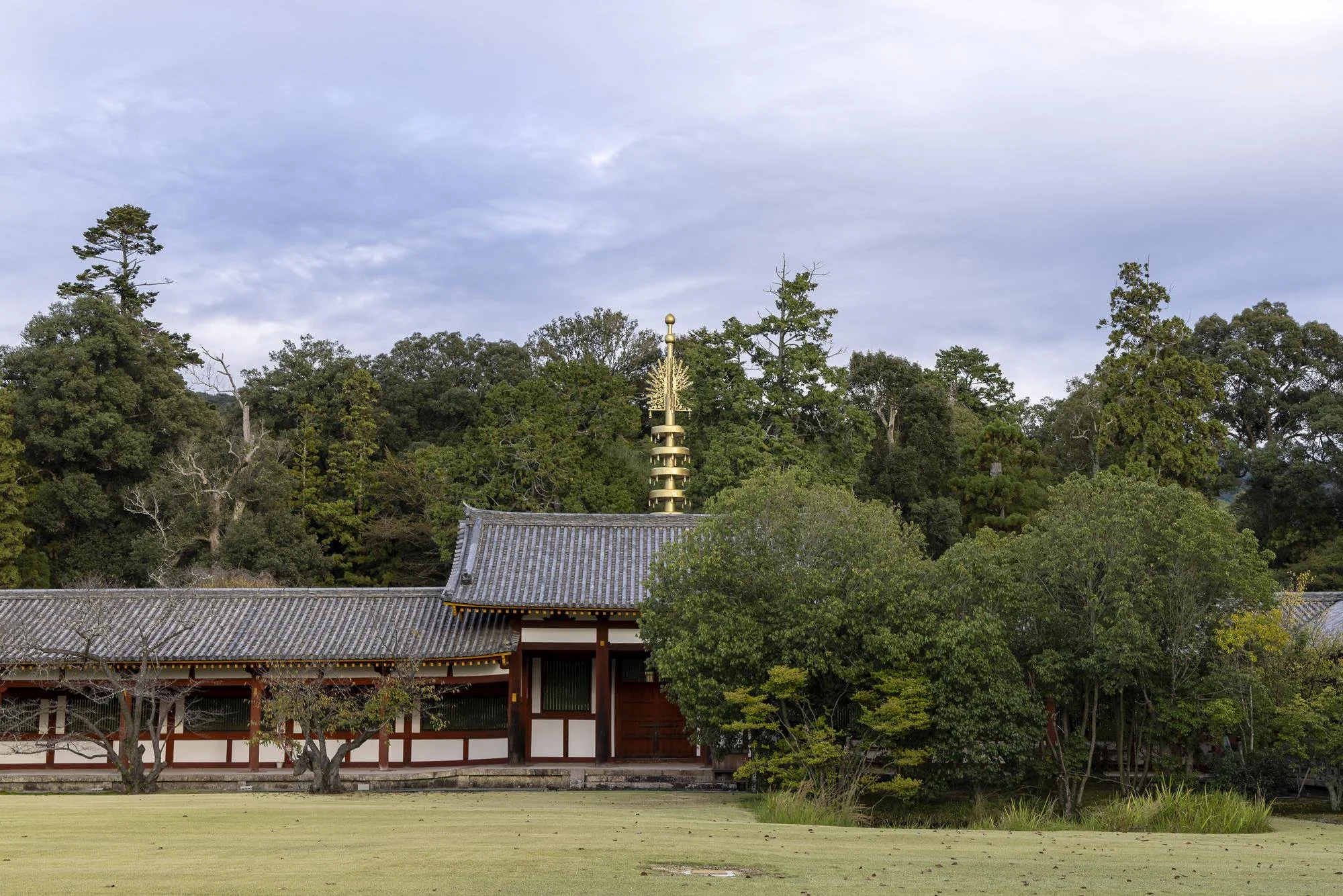 A traditional Japanese temple with a wooden structure, surrounded by trees and a grassy area, under a cloudy sky.