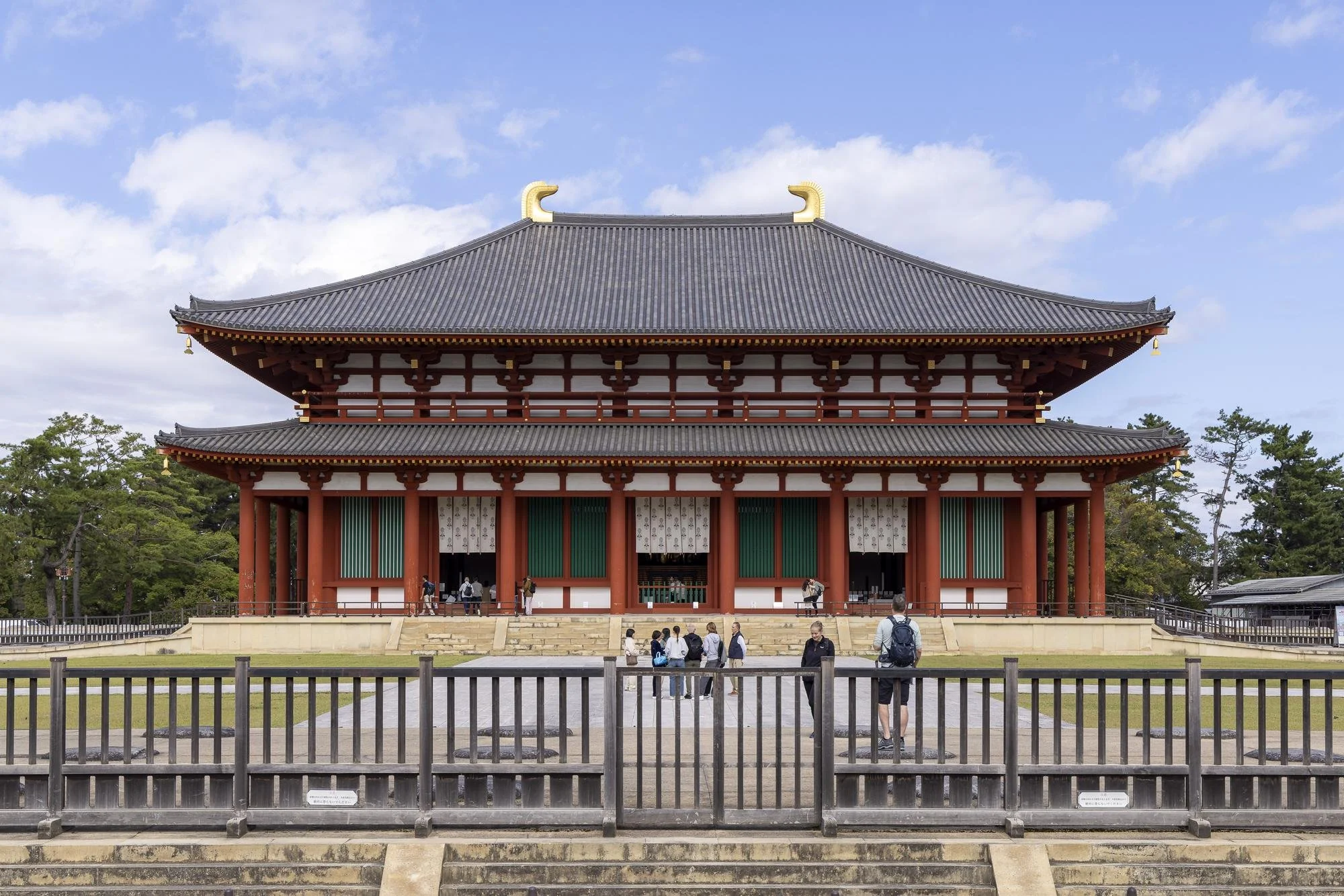 Kofuku-ji Temple: A traditional Japanese pagoda-style building with multiple tiers, red and white exterior, dark tiled roof, and decorative elements, with visitors walking on the steps and surrounding area under a partly cloudy sky.