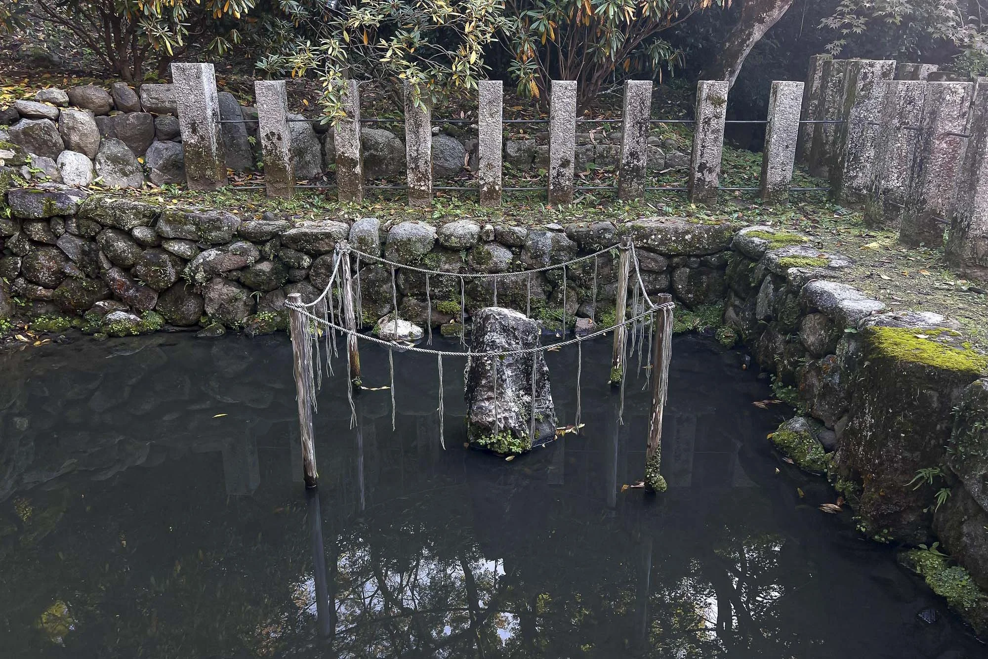 Small pond surrounded by stone wall and wooden fence, with moss-covered stones and trees in the background.