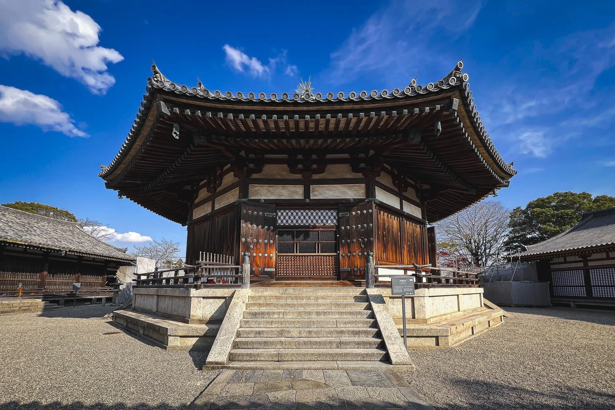 A traditional Japanese wooden building with a curved, ornate roof, accessed by stone steps, under a blue sky with a few clouds.