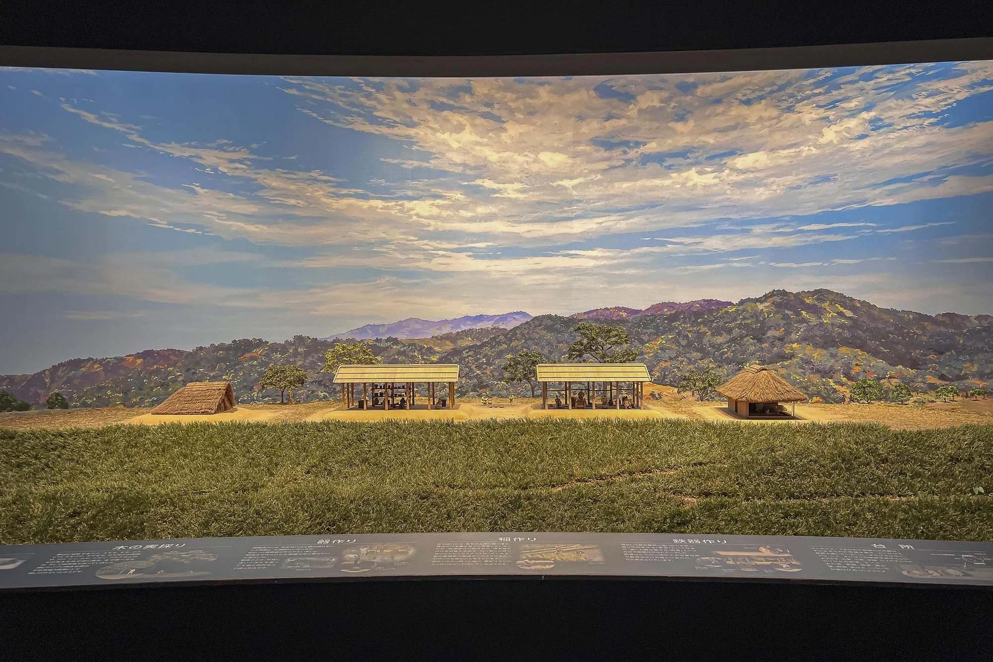 A museum display showing a miniature or model of a traditional village scene with thatched huts, trees, and mountains under a partly cloudy sky.
