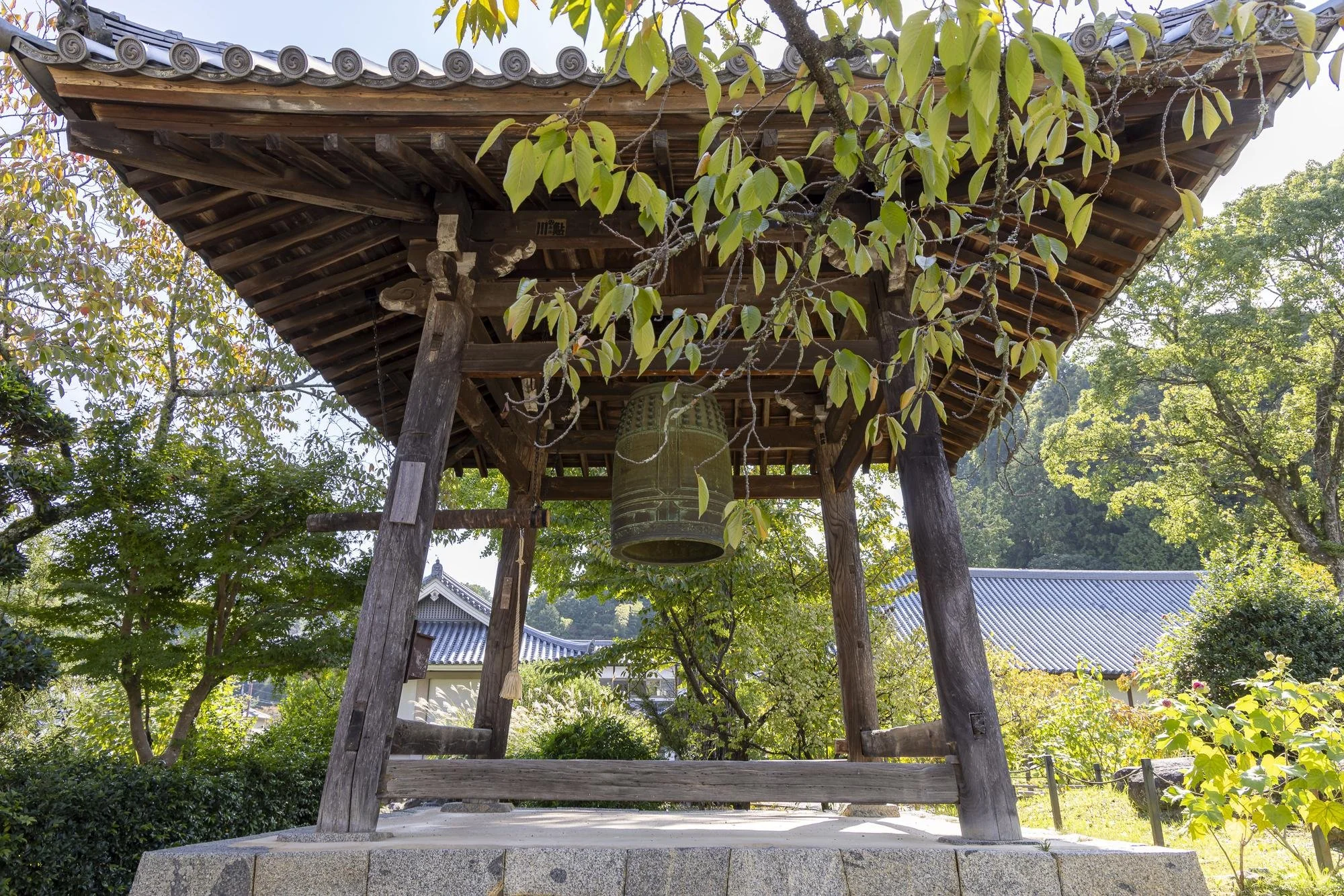 Traditional wooden bell tower with a hanging bronze bell, surrounded by green trees and foliage, in a Japanese garden setting.