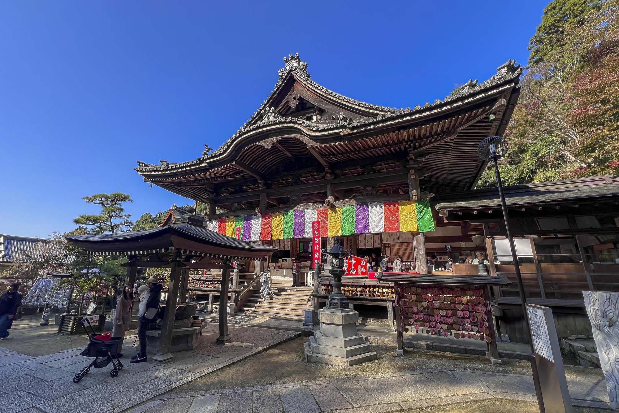 A traditional Japanese shrine with a wooden structure and ornate roof, decorated with colorful banners. Visitors are seen walking and praying around the shrine on a clear, sunny day.