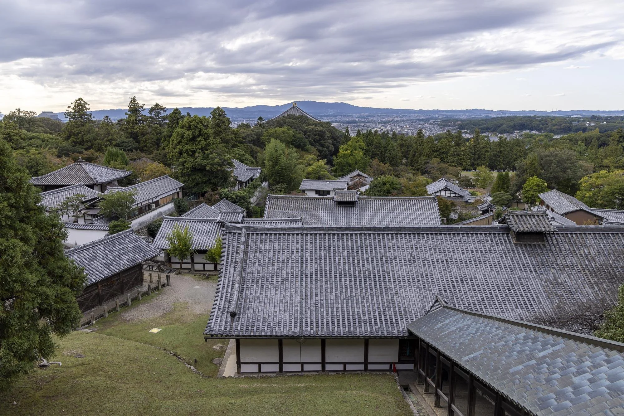 A traditional Japanese temple complex with tiled roofs, surrounded by trees and greenery, with a cityscape and mountains in the background.