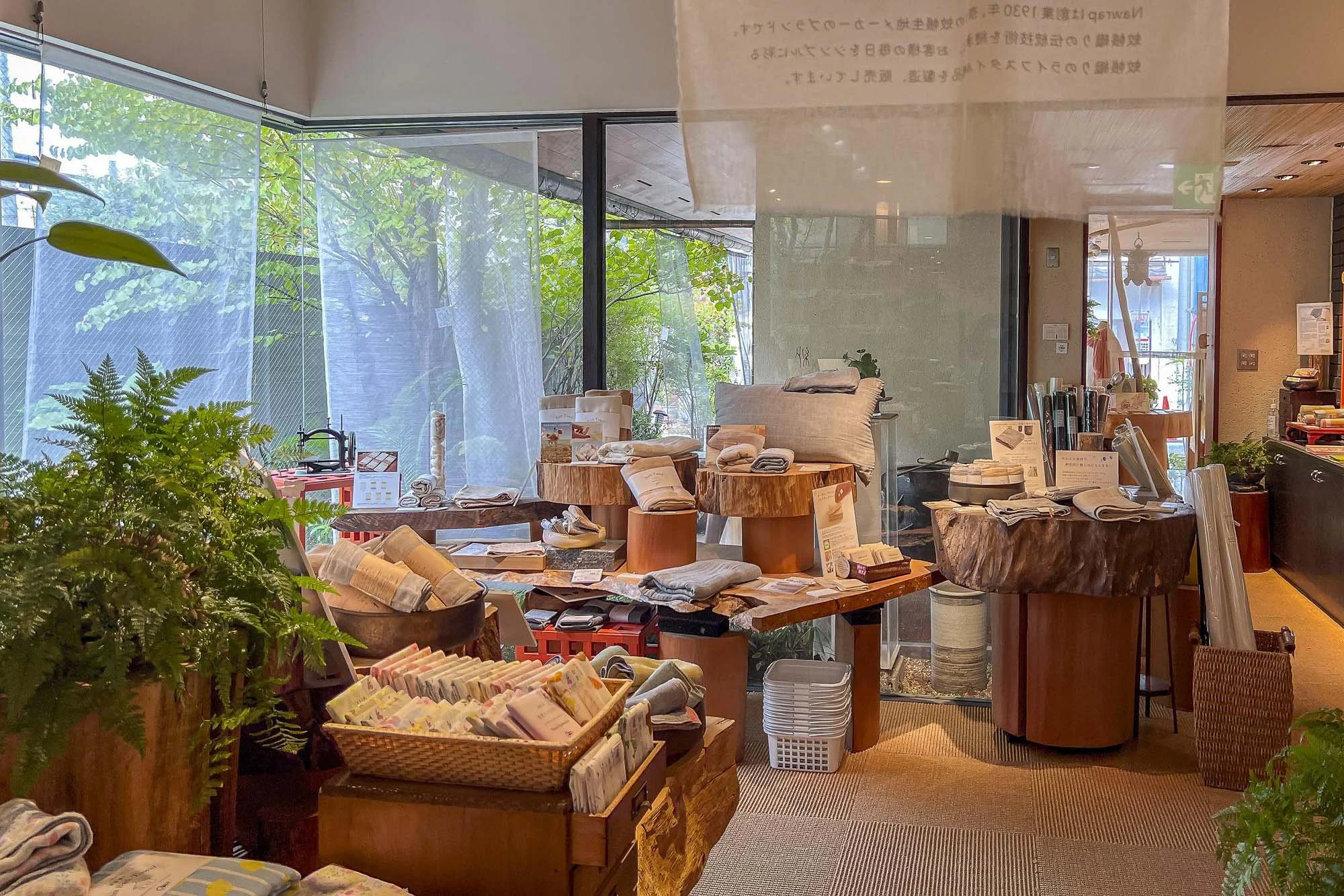 Display of home textiles and decor items inside a retail store, with large windows showing green trees outside.