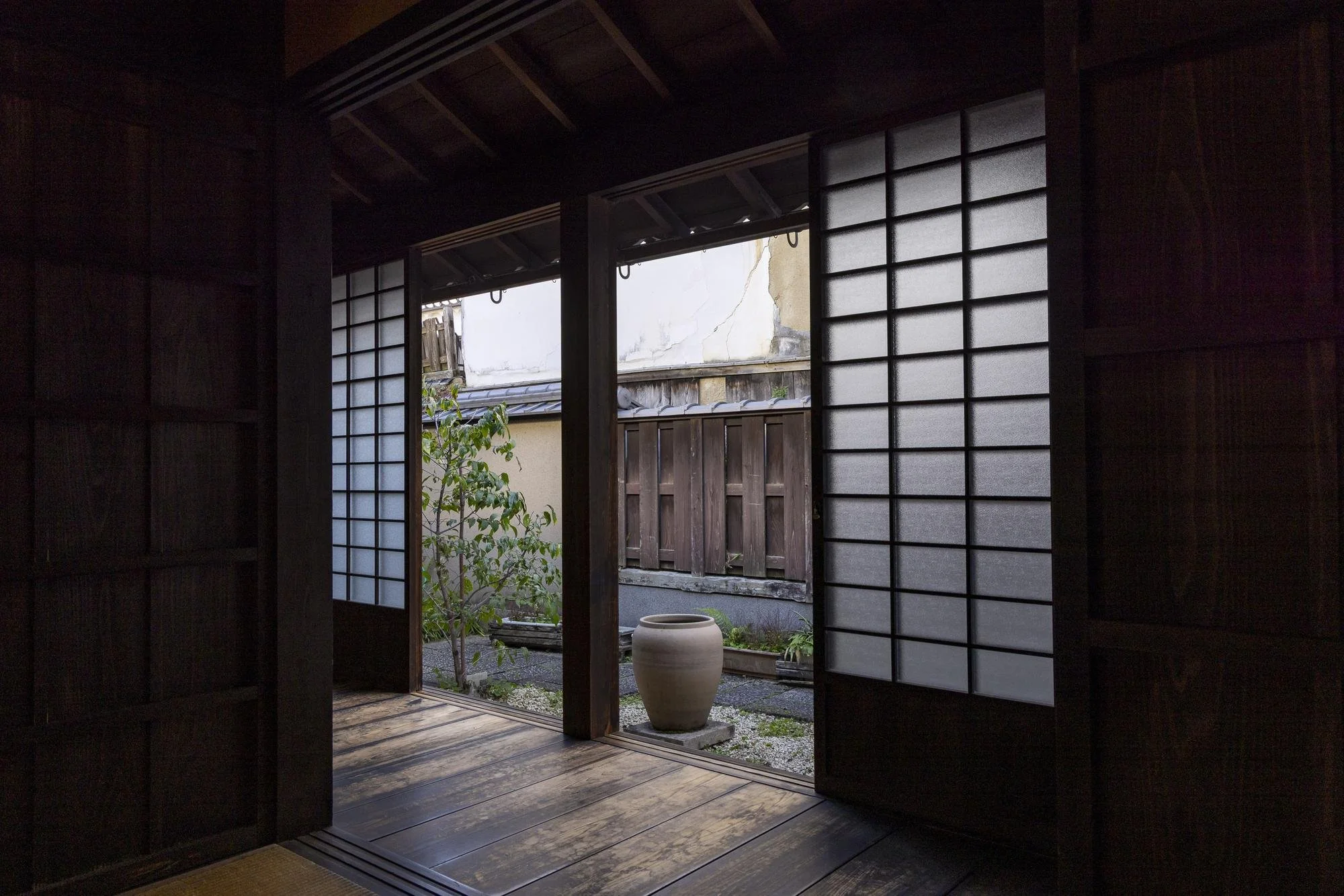 Traditional Japanese shoji doors open to a small outdoor garden with a large ceramic pot, a tree, and a wooden fence.
