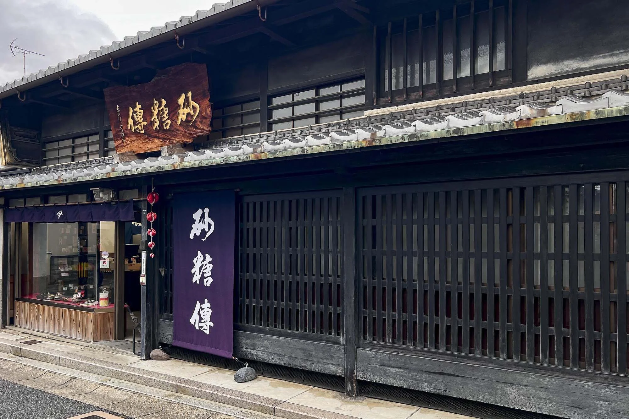 Traditional Japanese building with dark wood slats, purple banner with white Japanese characters, and a wooden sign with gold Japanese characters, featuring a display window and small decorative red lanterns.