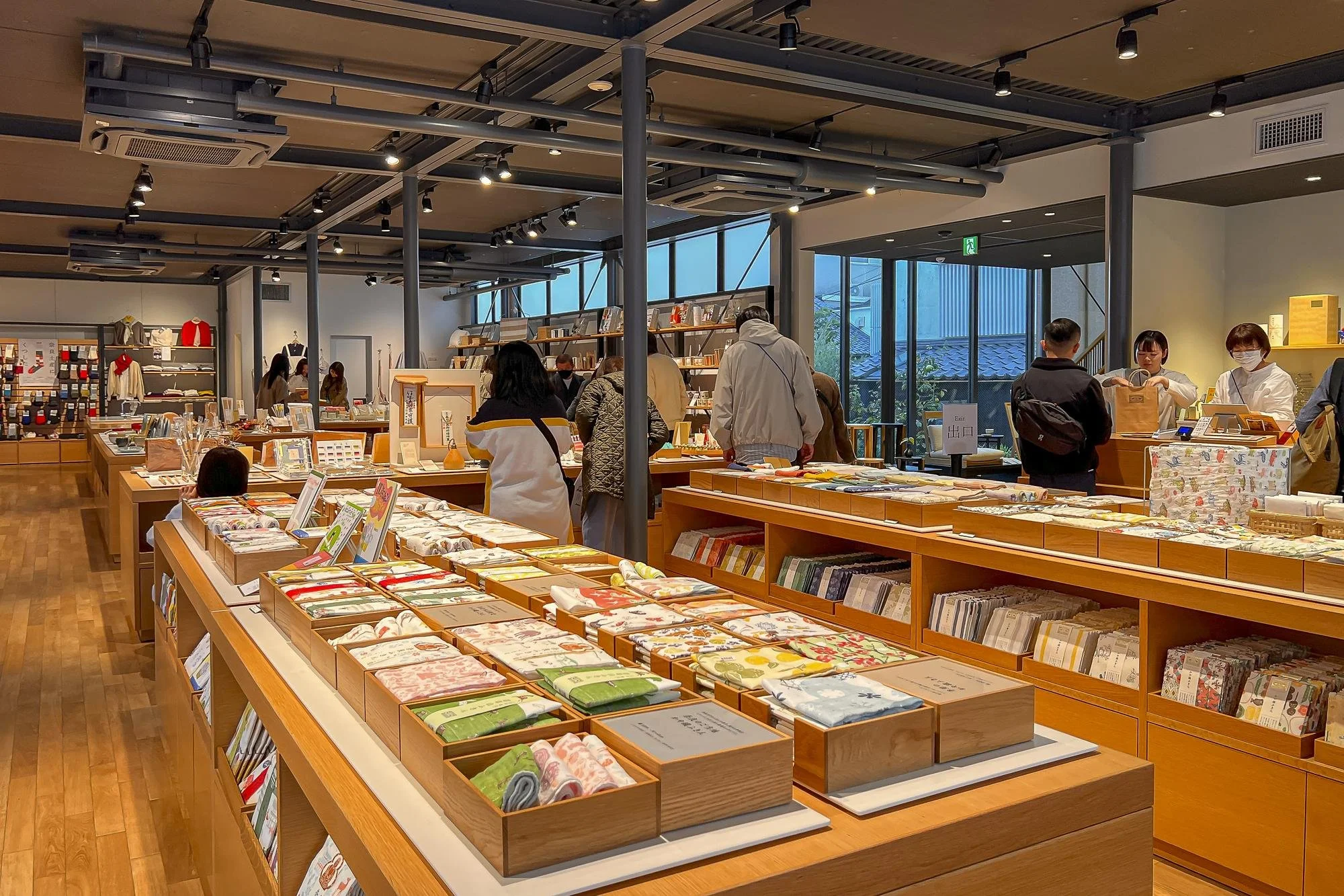 Interior of a store with wooden display tables holding fabric items, with customers browsing and employees assisting, and large windows in the background.
