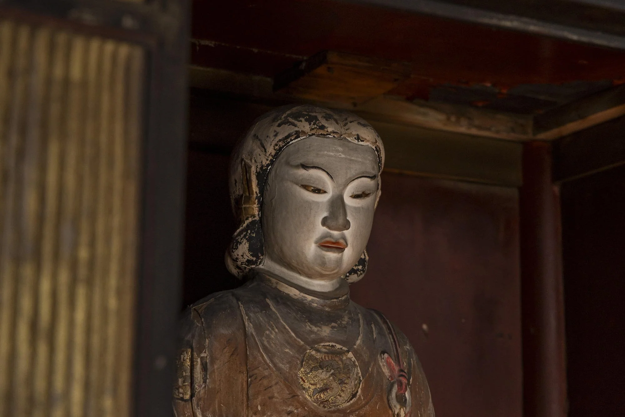 An antique Asian-style wooden sculpture of a woman's face and upper body, partially concealed behind wooden slats, with a wooden dark brown background and ceiling.