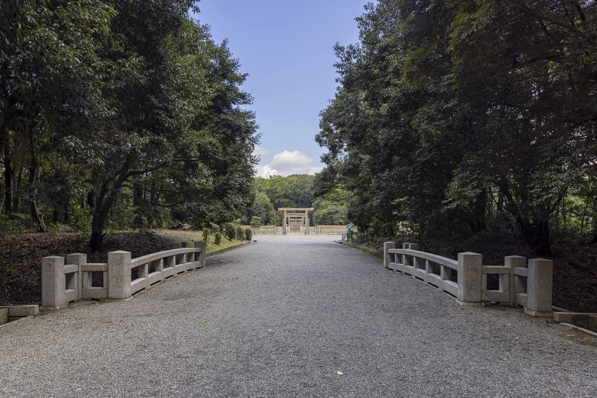 A gravel pathway leading to a traditional shrines or temple gates, framed by lush green trees on both sides.