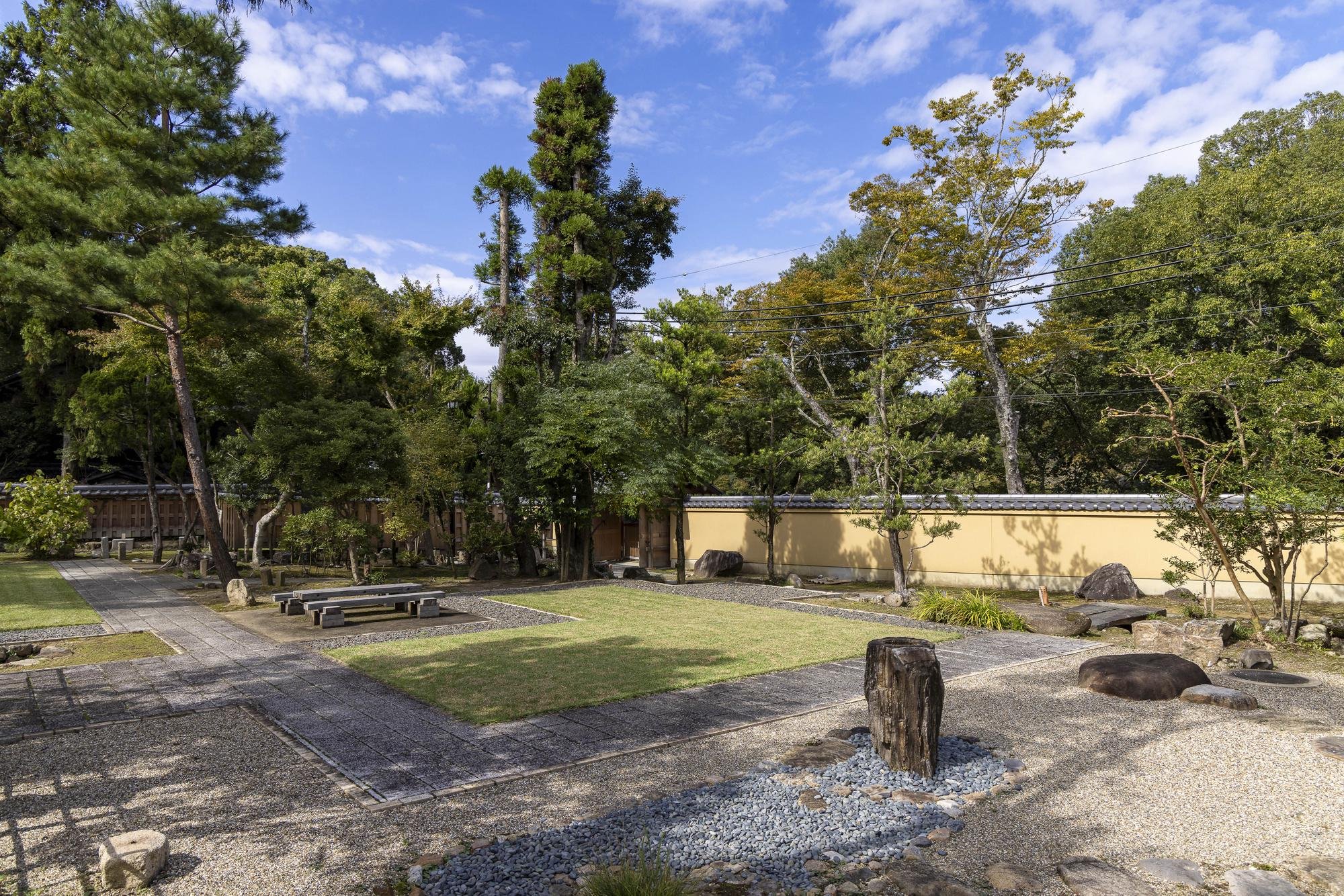 A traditional Japanese garden with a stone pathway, benches, manicured grass, rocks, and trees, enclosed by a yellow wall and greenery, under a partly cloudy sky.