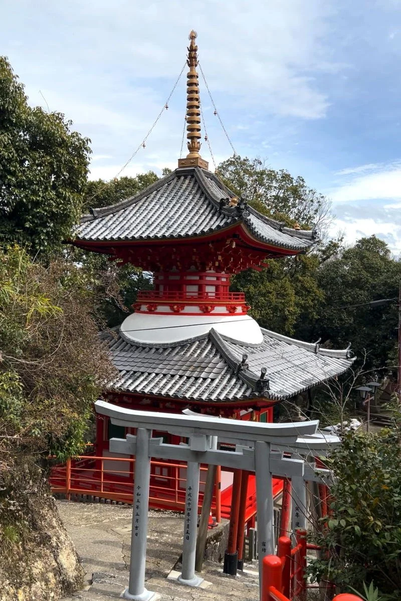 A traditional Japanese pagoda with multi-tiered roofs, a tall ornate spire on top, and a torii gate at the base, surrounded by trees and a stone pathway.