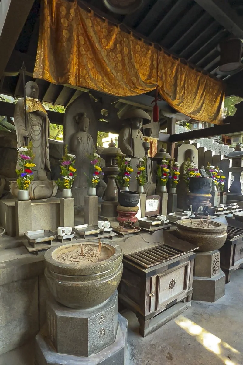 Buddhist altar with stone statues, candles, incense, and colorful flowers under a golden curtain.