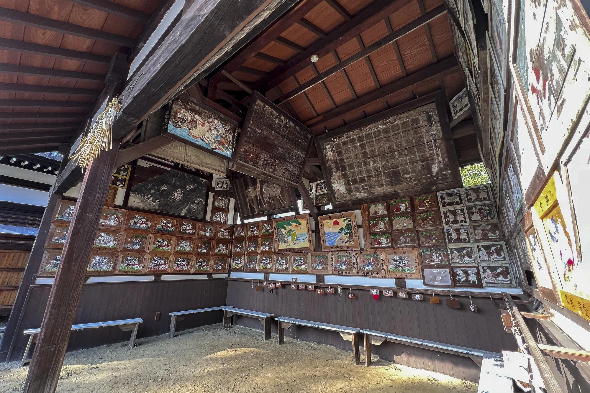 Interior of a traditional Japanese shrine or temple with wooden panels and colorful artwork, including many small paintings and some hanging ornaments.