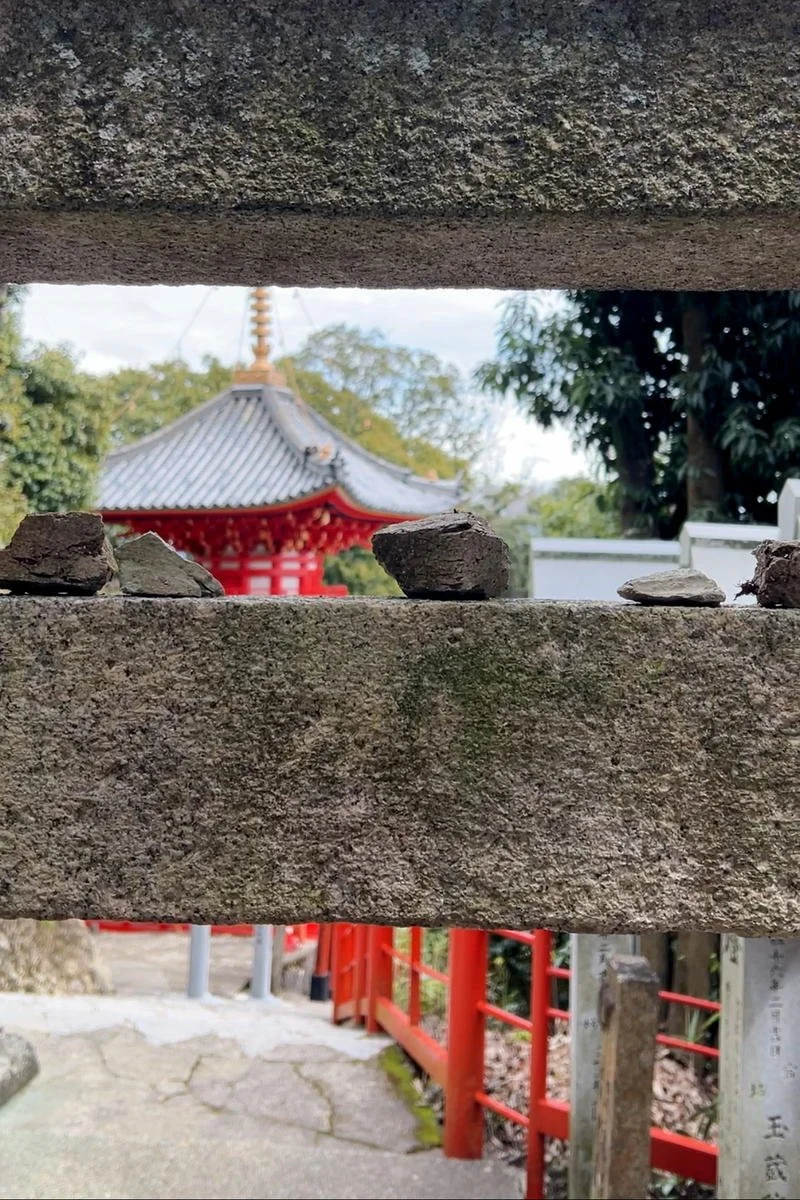 A photograph taken from behind a stone wall with small rocks on top, showing a traditional Japanese temple or pagoda with a gray, curved roof and red structure in the background, surrounded by green trees and a partly cloudy sky.