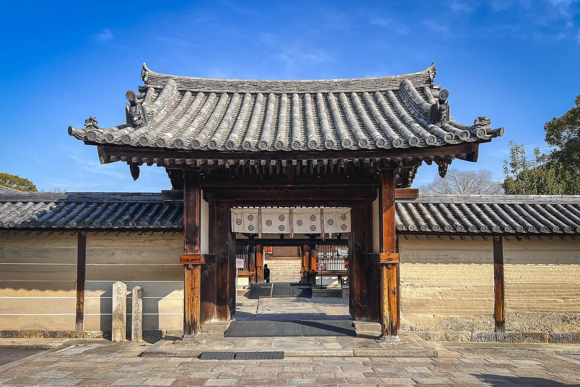 Traditional Japanese temple gate with wooden beams, tiled roof, and a stone wall under a blue sky.