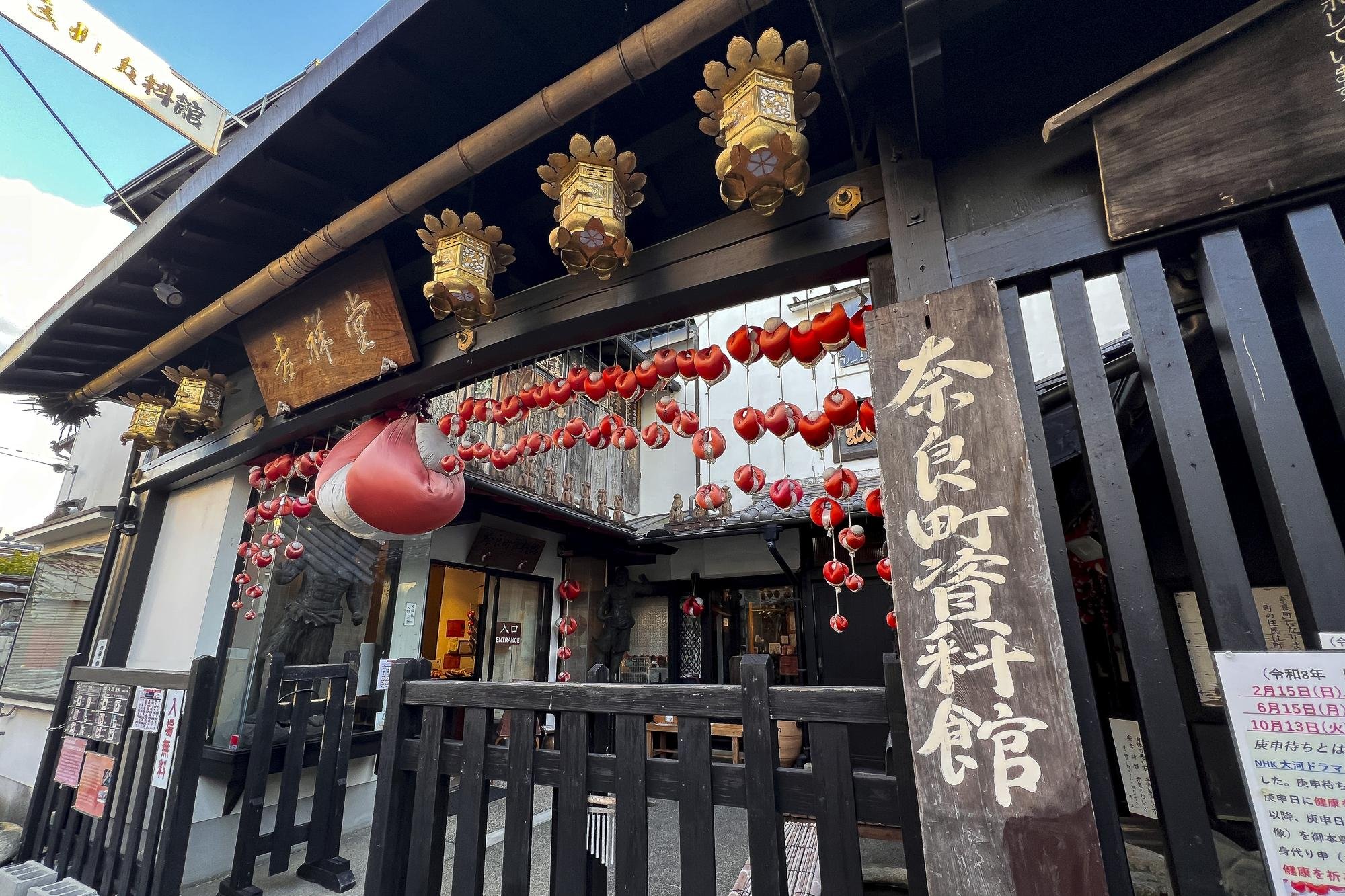 Exterior of a Japanese shrine or temple decorated with red lanterns and a large paper lantern, with a wooden sign in Japanese characters and hanging lanterns.