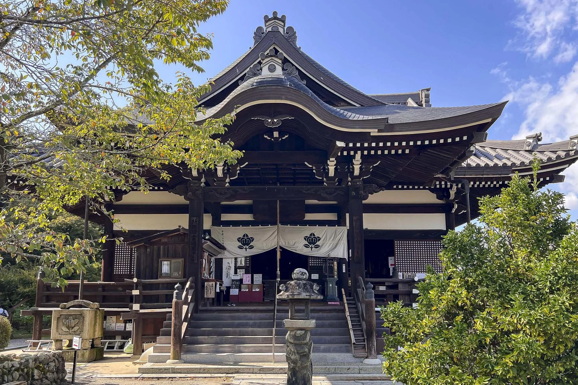 A traditional Japanese shrine with a wooden structure, curved roof, and steps leading to the entrance, surrounded by greenery and trees under a blue sky.