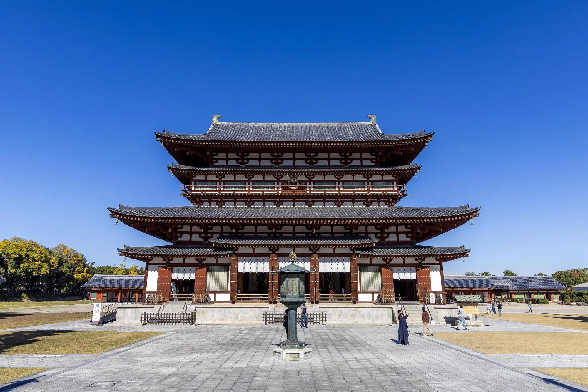 A traditional Japanese multi-tiered wooden temple building under a clear blue sky, with a stone path and lantern in the foreground and small groups of people walking around.