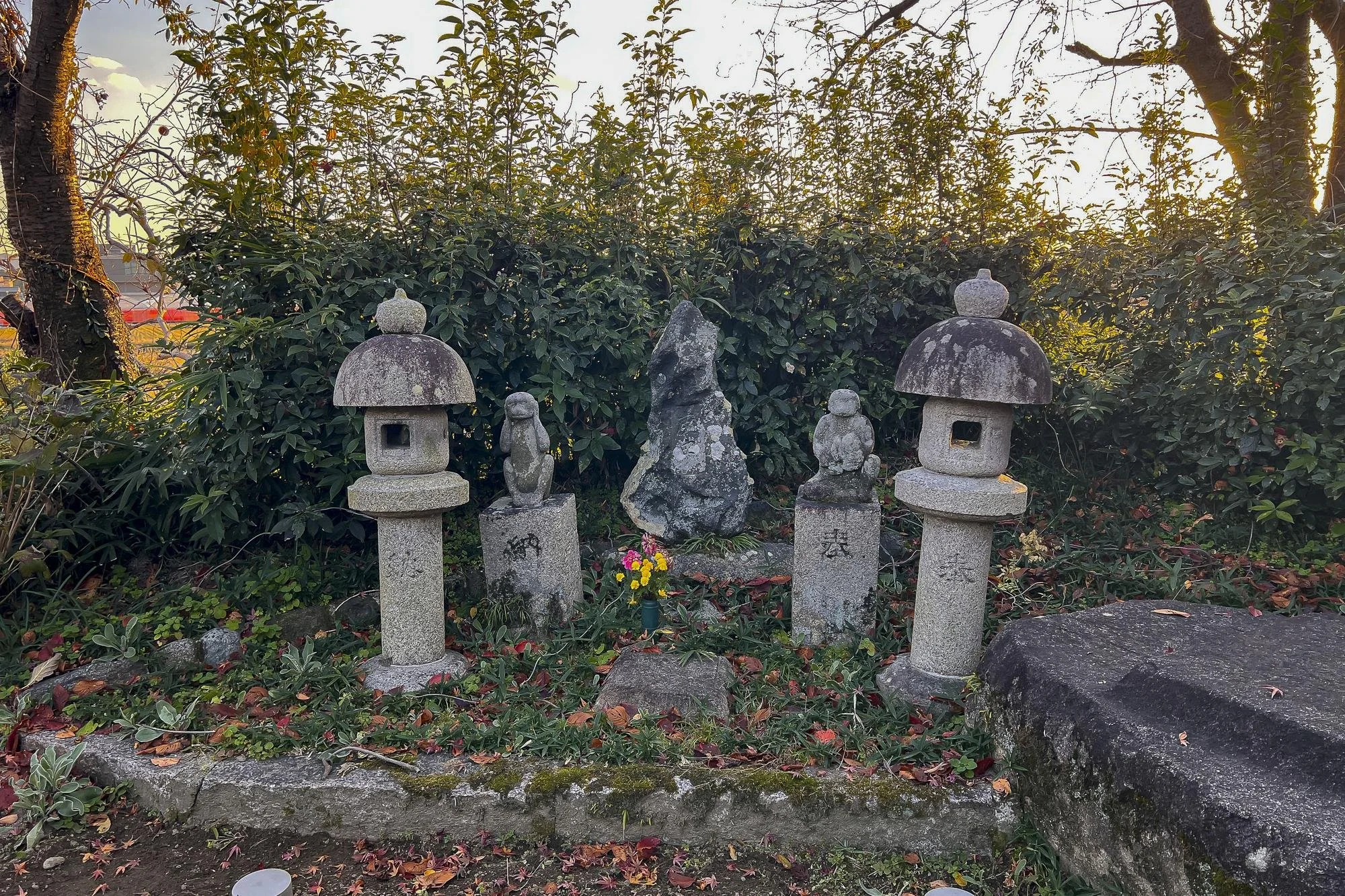 A small Japanese-style garden shrine area with stone lanterns, statues, and a rock centerpiece, surrounded by greenery and fallen leaves.