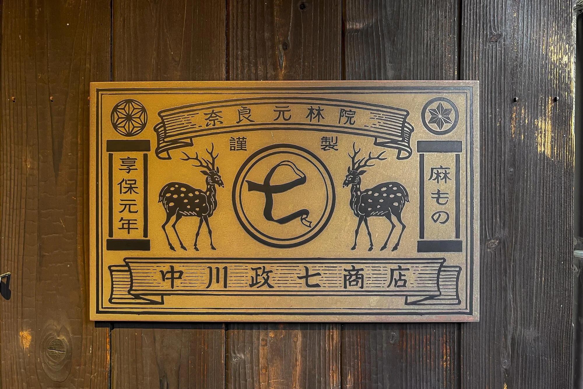 A decorative wooden sign with Japanese characters, two illustrated deer with antlers, and symbols, mounted on a dark wooden wall.
